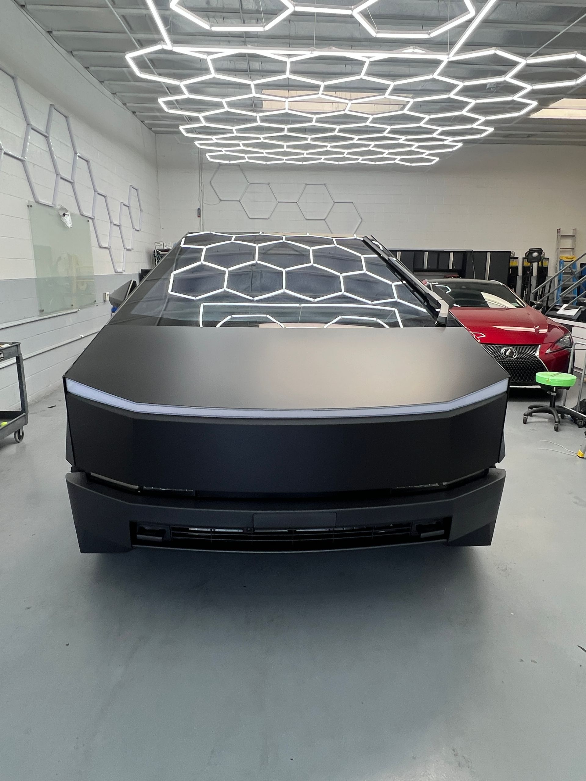 Tesla Cybertruck in a showroom, front view, with a matte gray body and geometric ceiling lights reflected on the windshield.