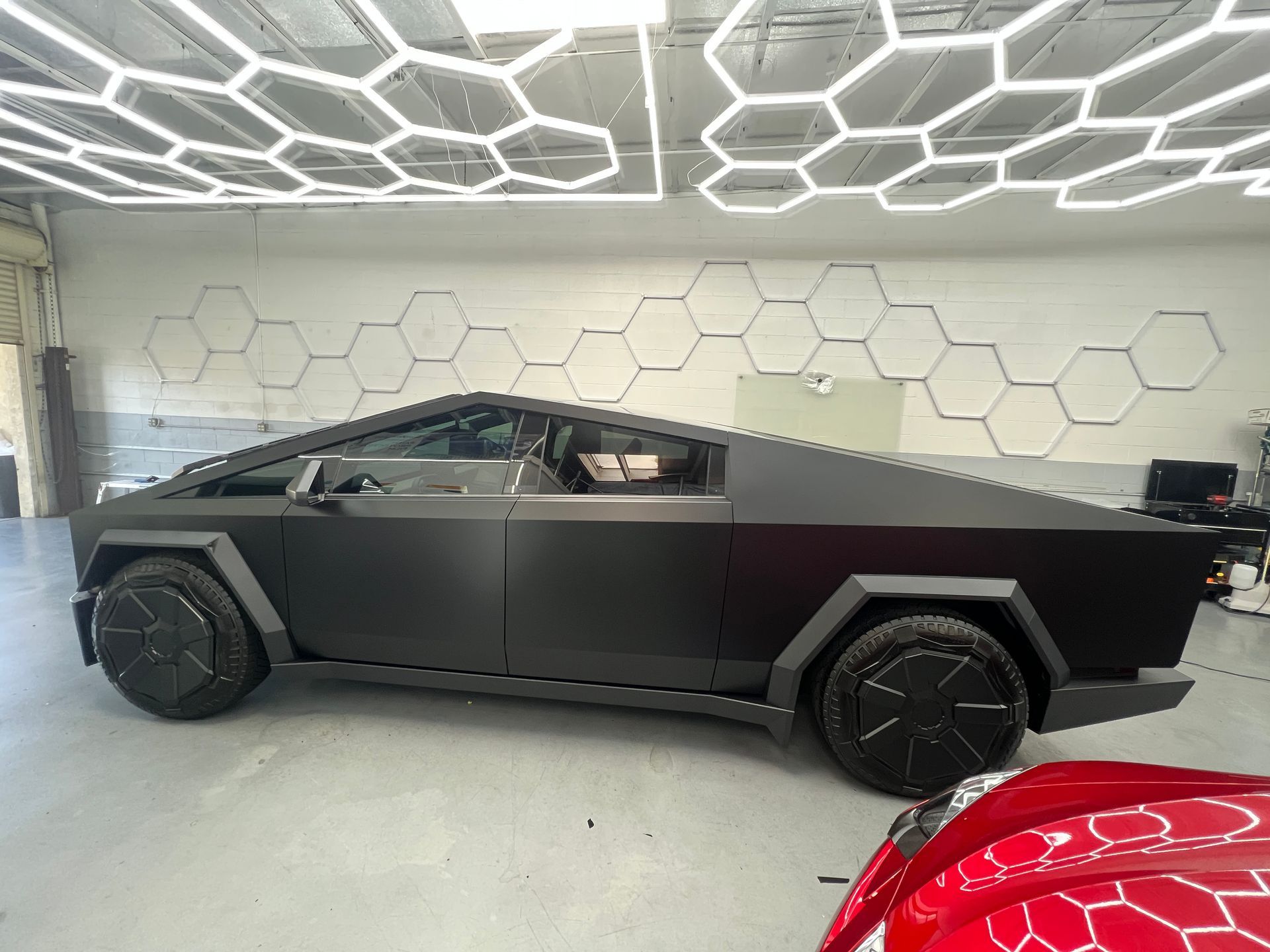 Black Tesla Cybertruck indoors under hexagonal ceiling lights, with a red car partially visible in front