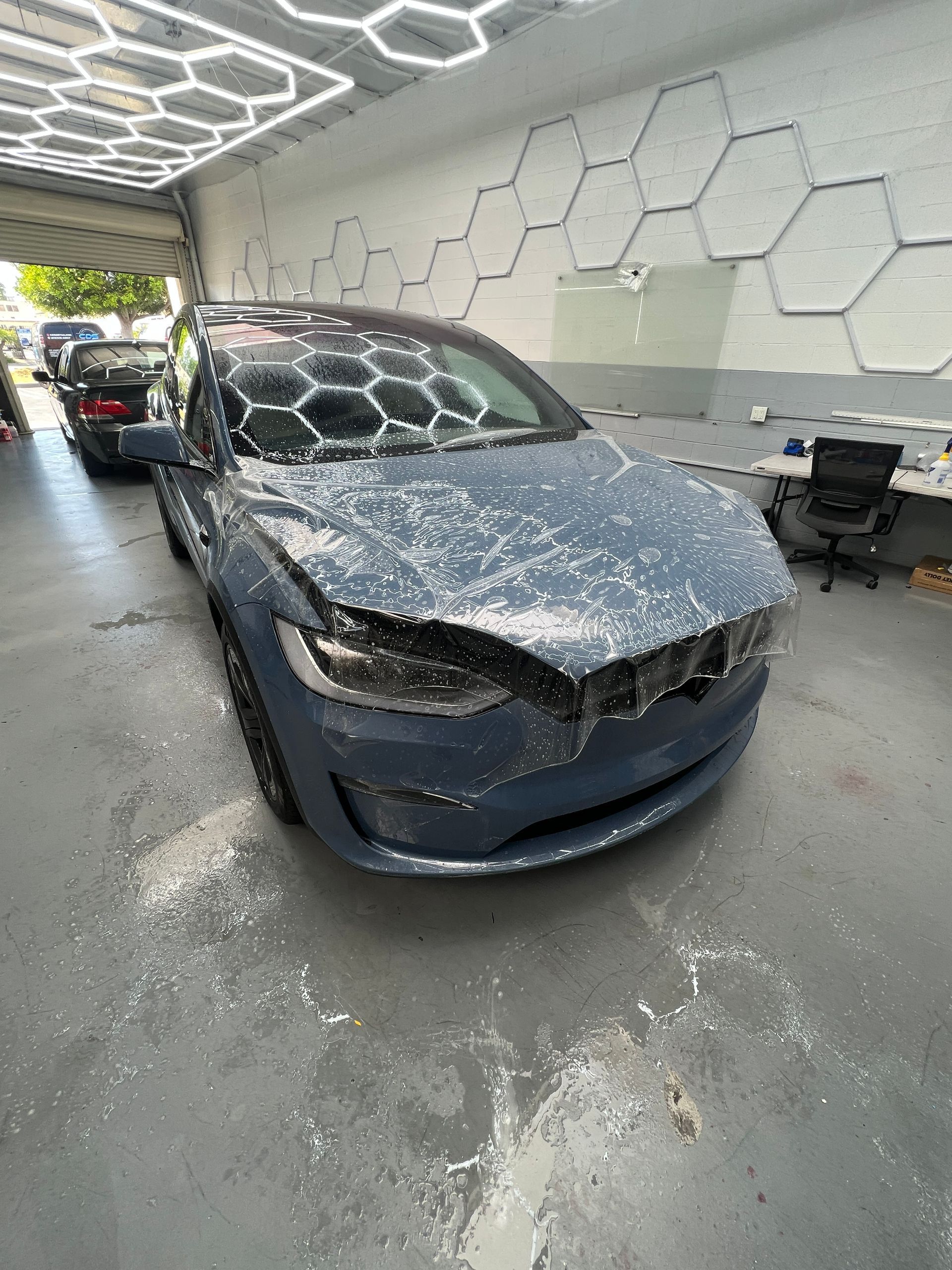 Blue Tesla sedan with a cracked windshield inside an auto body shop, under bright hexagon ceiling lights