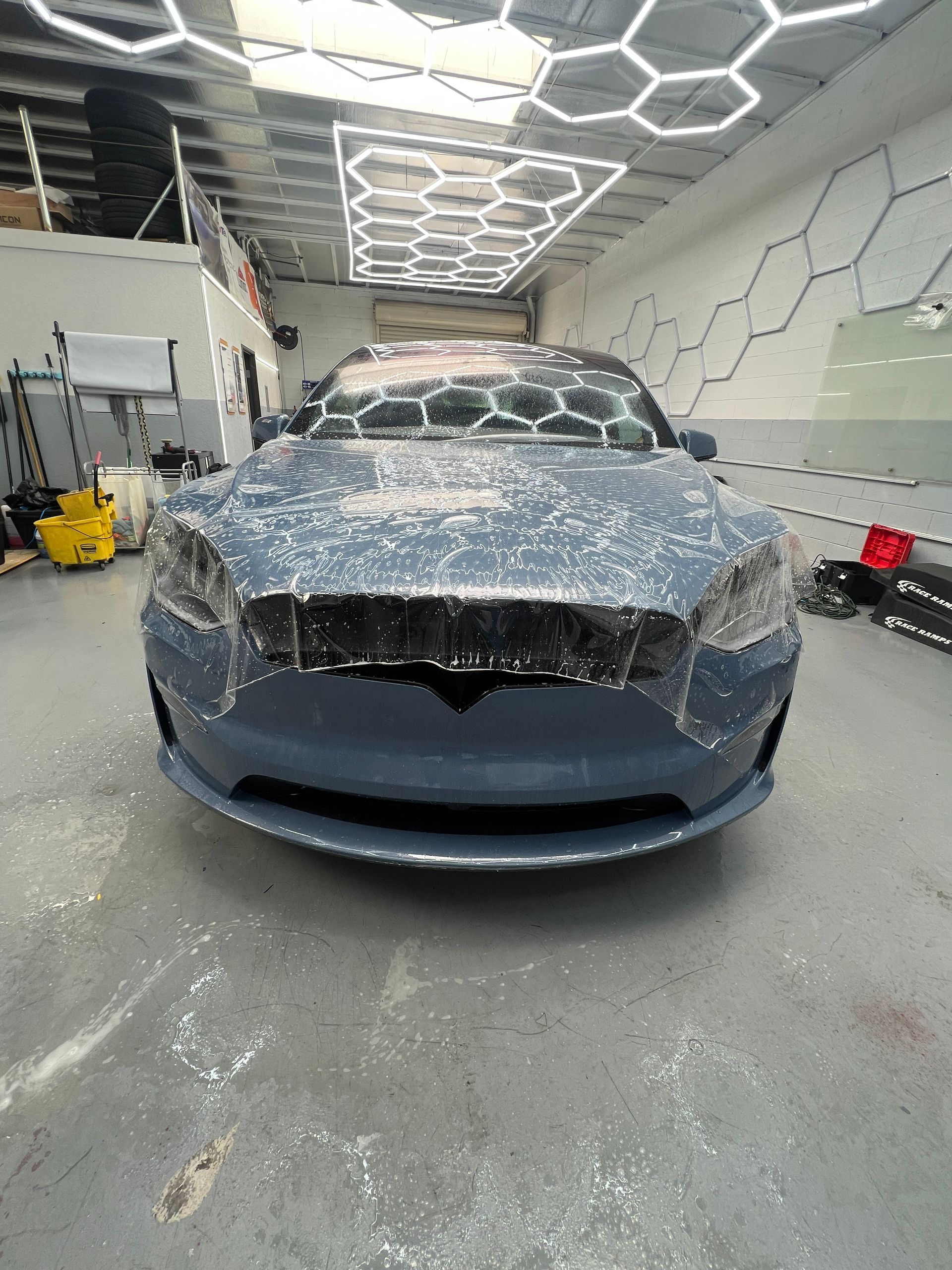 Blue car in an auto body shop with a dusty hood and front bumper removed under bright hexagon ceiling lights
