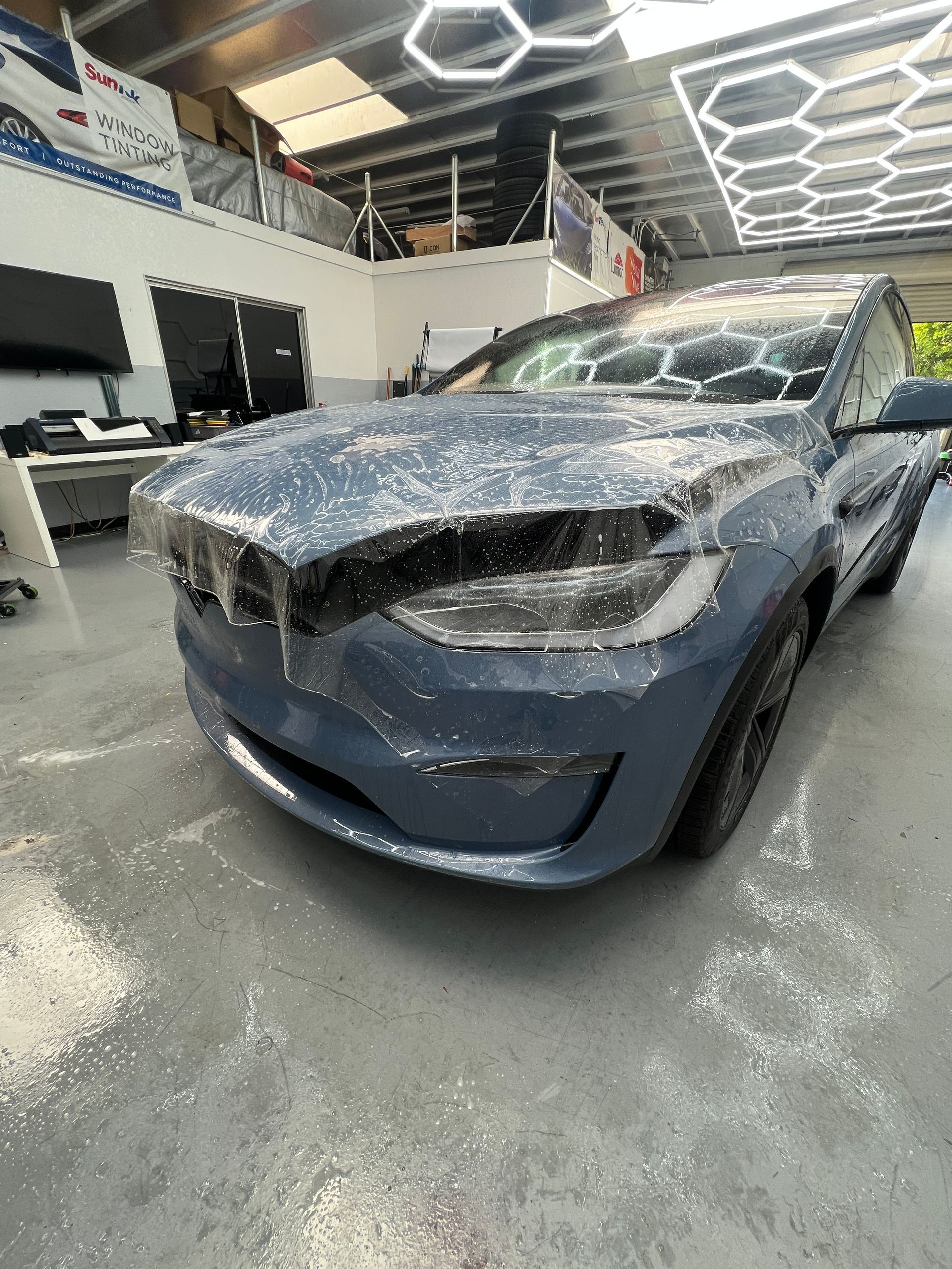Blue sedan wrapped in plastic film inside an auto shop with bright hexagon ceiling lights
