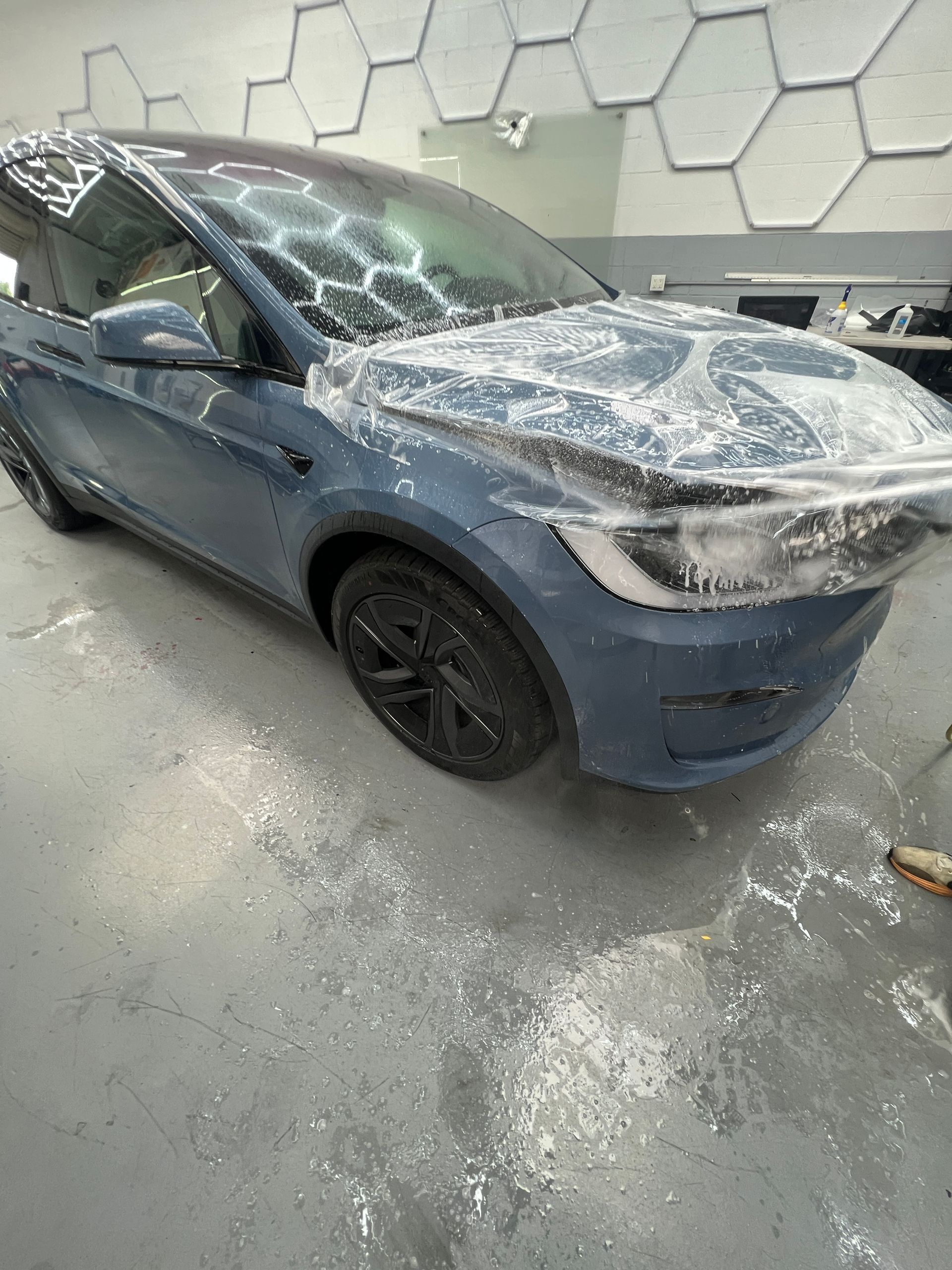 Blue sedan covered in a translucent protective film in a workshop, with black wheels and a wet concrete floor
