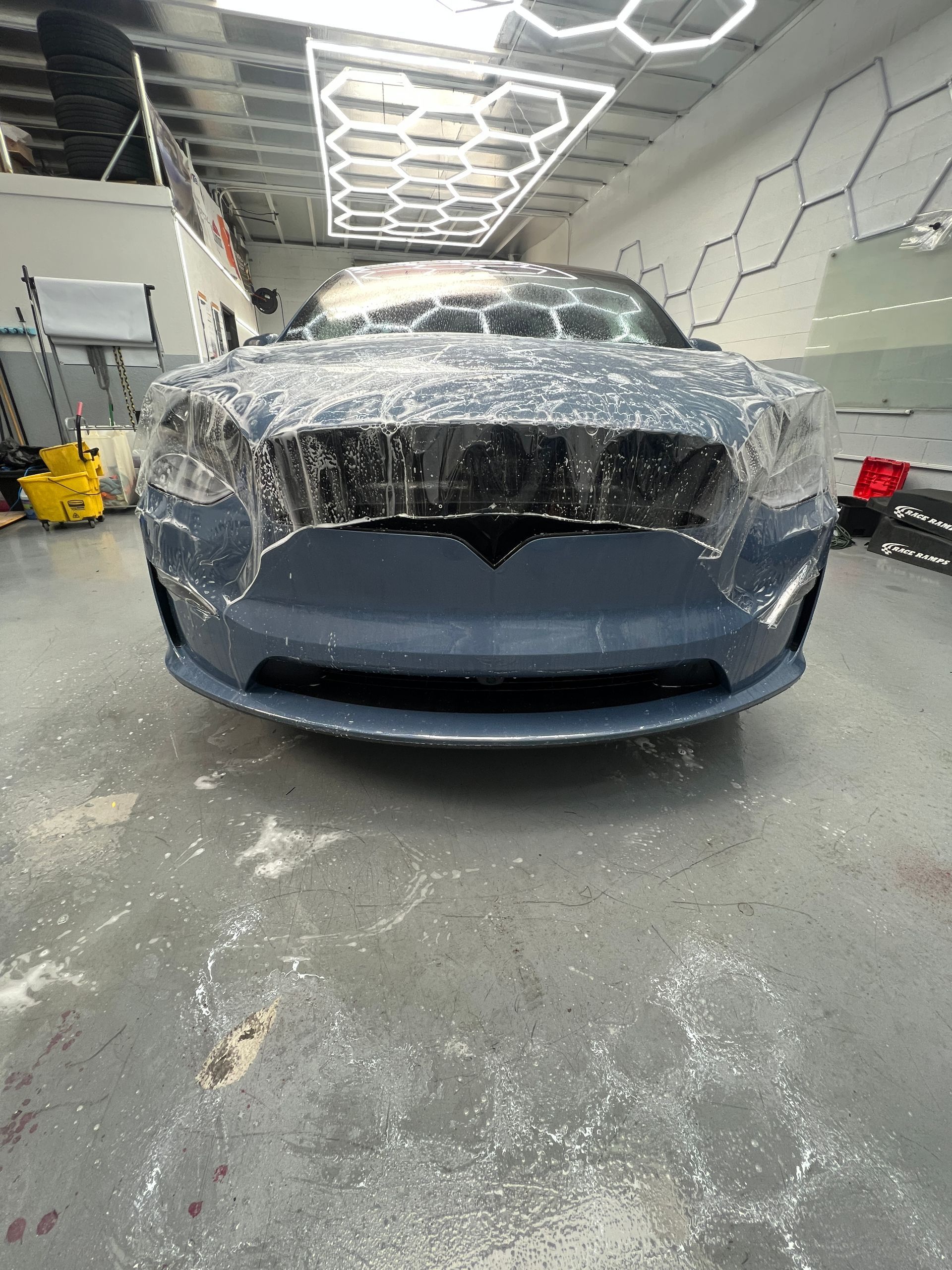 Front view of a gray car wrapped in plastic inside a modern auto workshop.