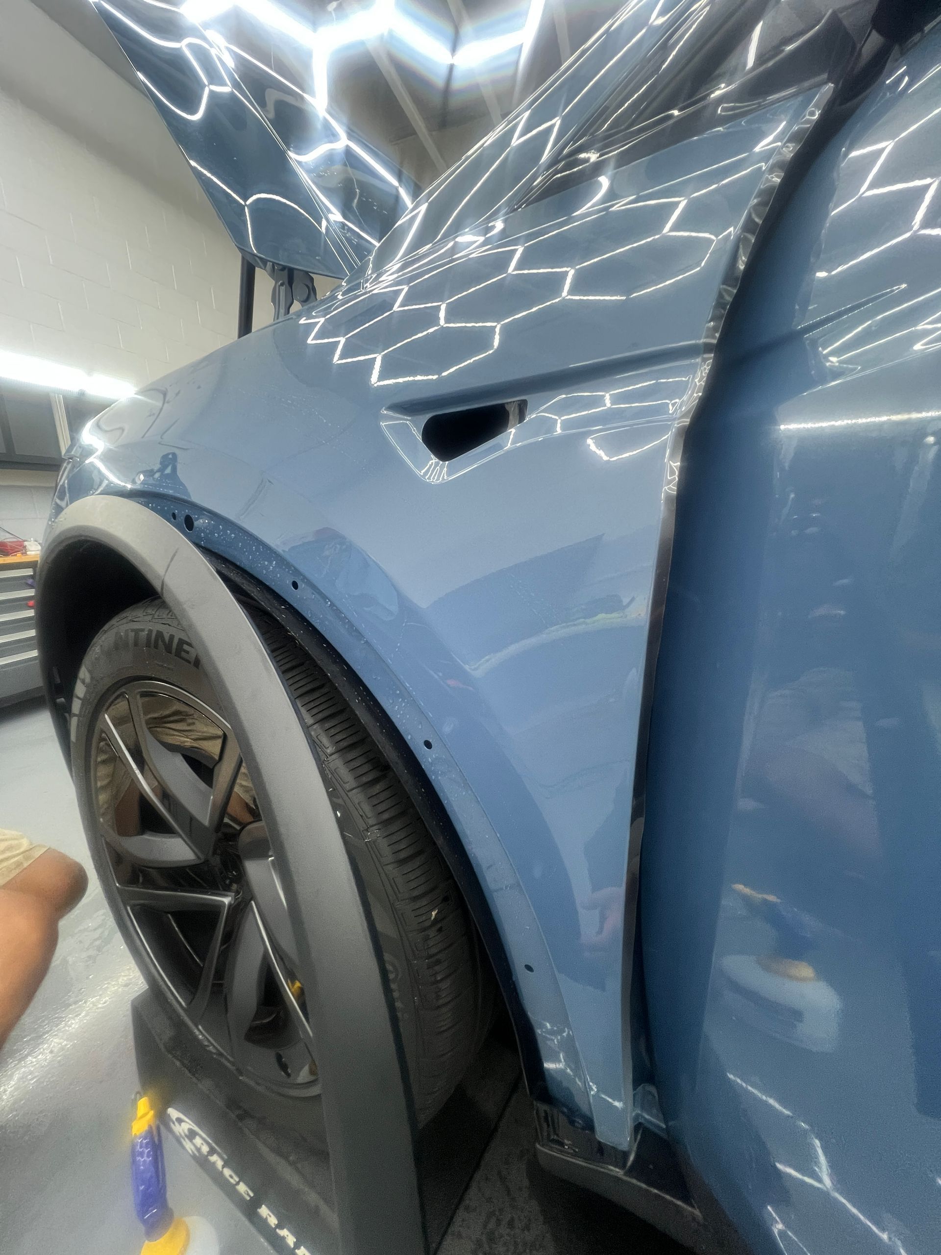 Blue car side panel and rear wheel under bright reflections, with a small dent near the fender