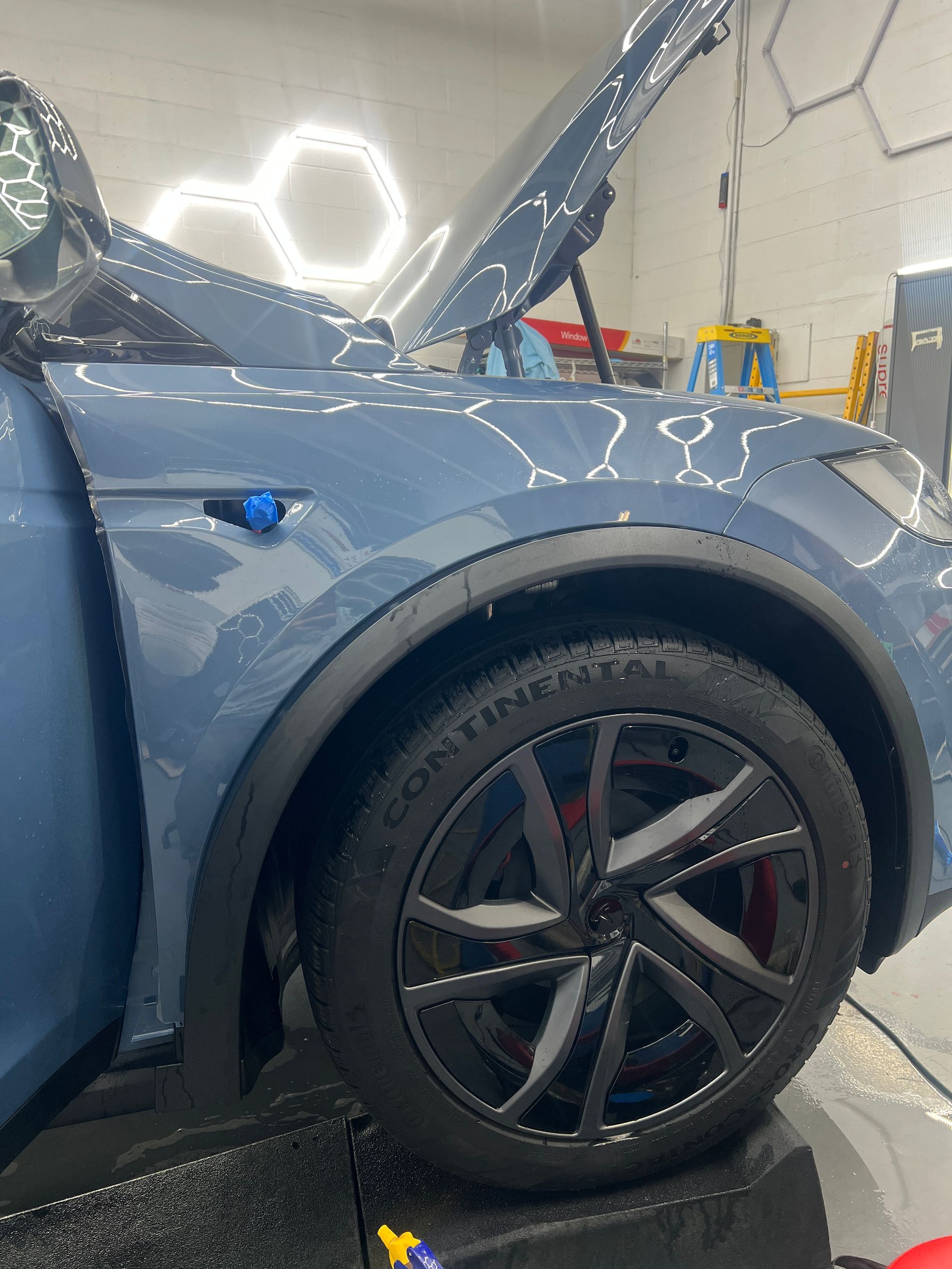 Blue car with raised hood in a workshop, showing the front wheel and fender.
