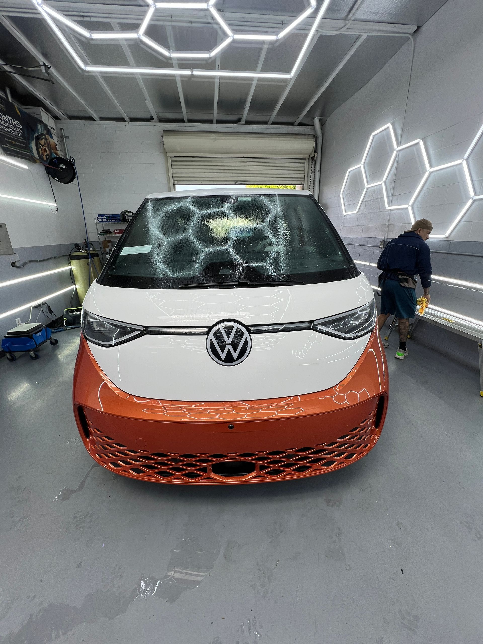 Volkswagen van in a workshop with orange front wrap and honeycomb wall lights; person working at right