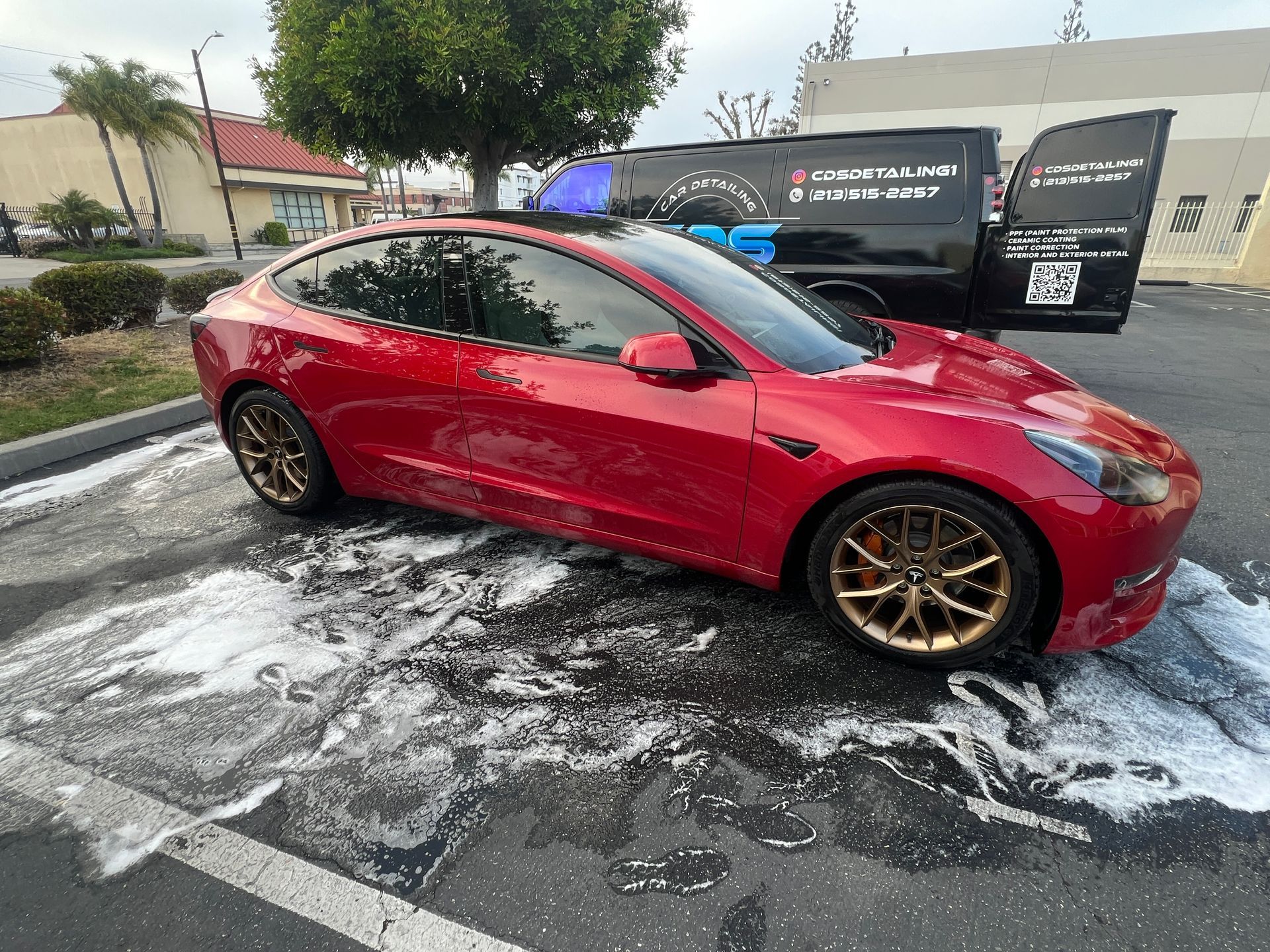 Red Tesla sedan parked on a wet, soapy lot near a mobile car detail trailer.