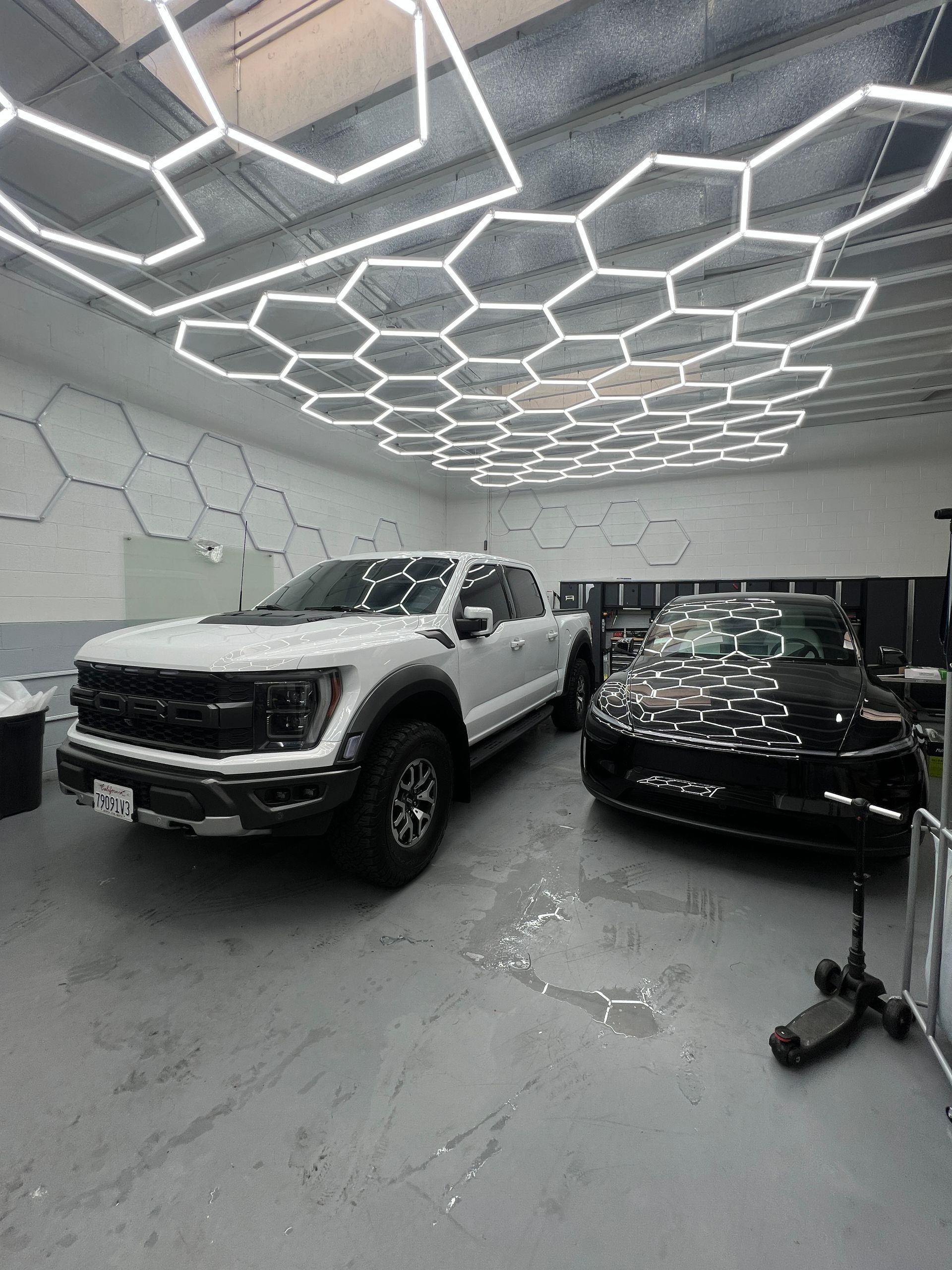 Two cars in a modern showroom under hexagonal ceiling lights, with a white pickup beside a black car.
