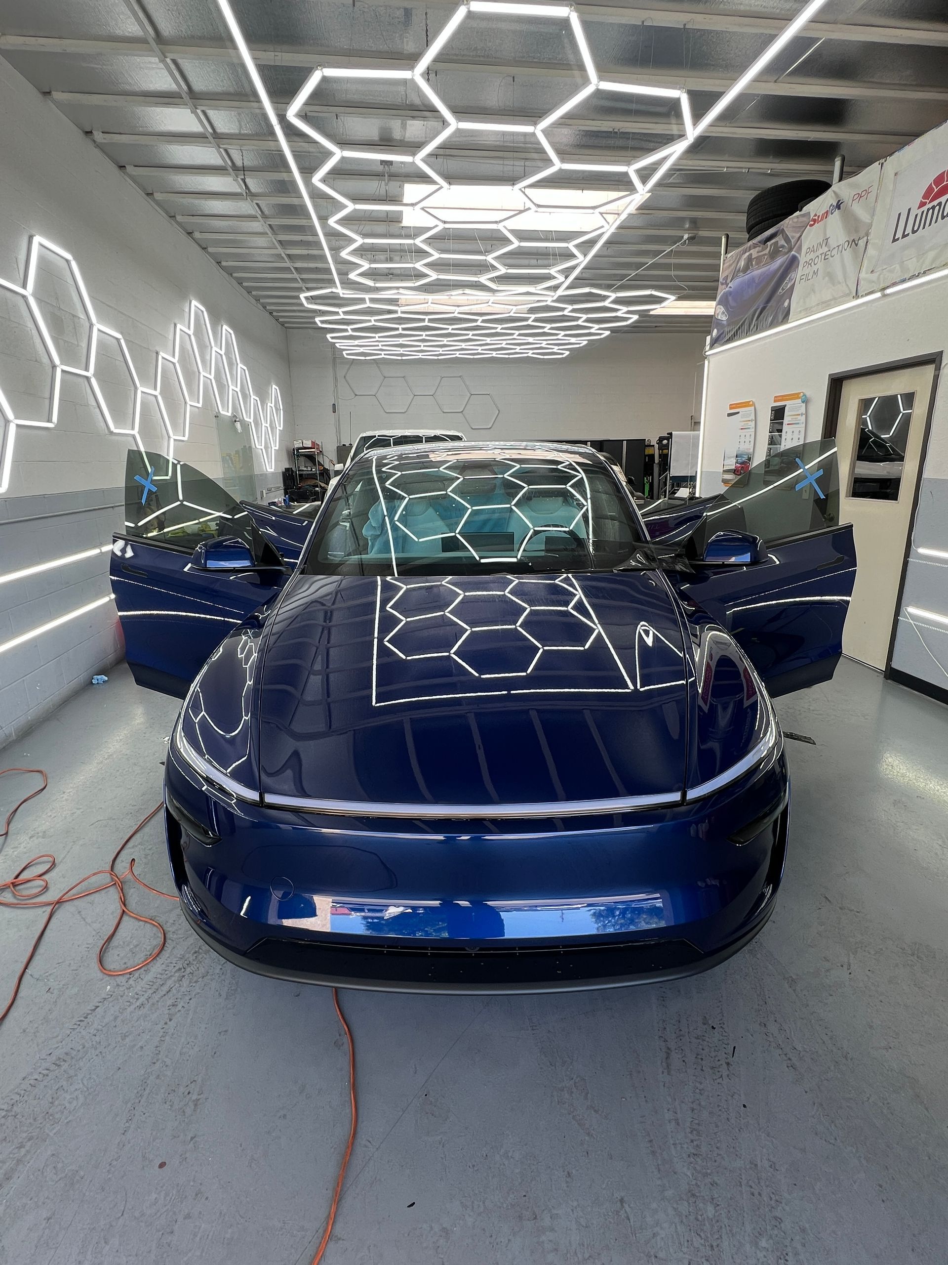 Blue sports car in a detailing bay with open doors and bright hexagonal ceiling lights reflected on the hood