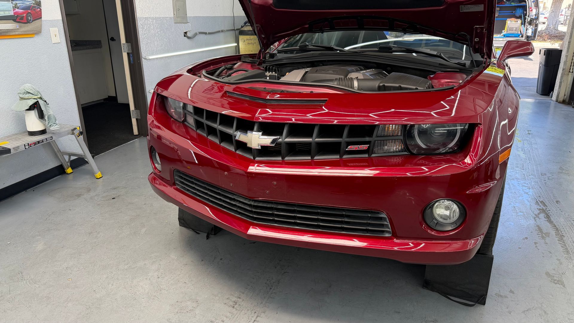 Red car in a garage with hood open, showing the front grille and headlights.