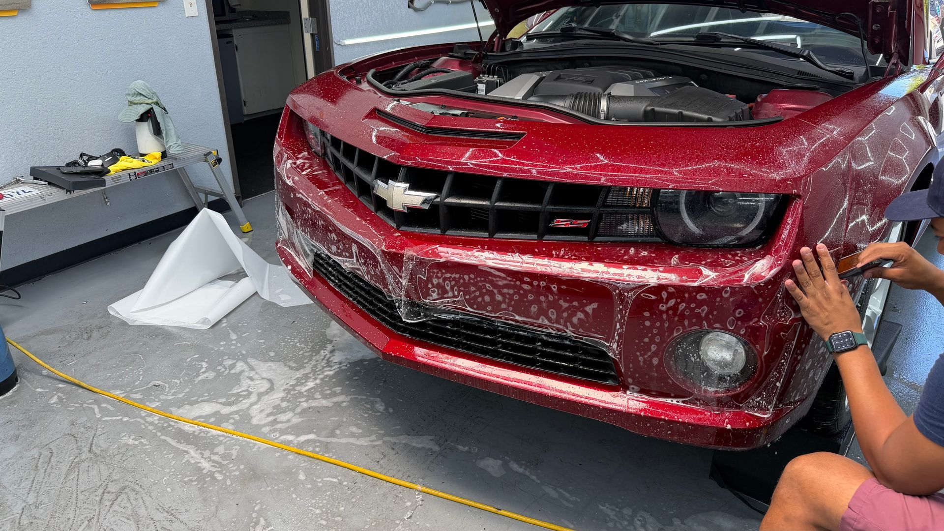 Red sports car being hand-washed in a garage, with soap suds and a person rinsing the front end.