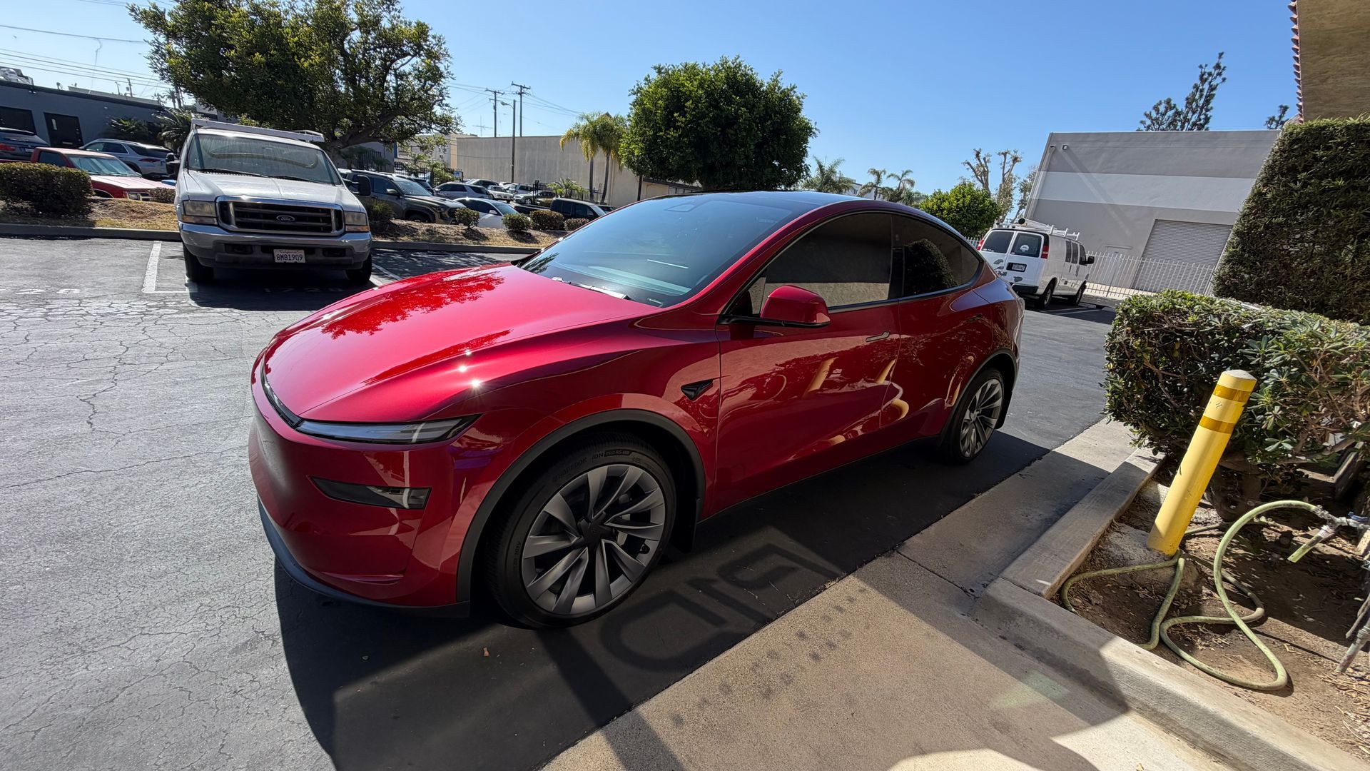 Red Tesla parked beside a curb in a sunny lot, with a white truck and yellow bollard nearby.