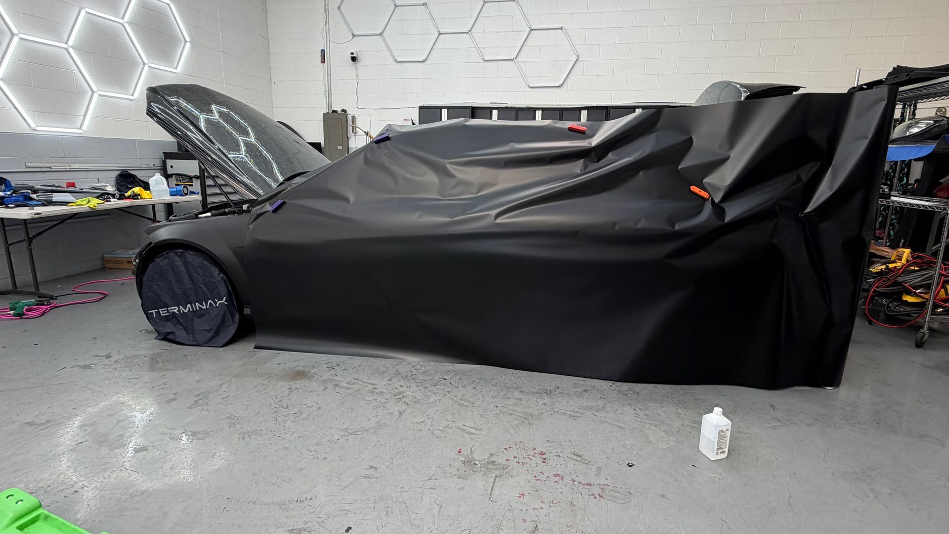 Black sports car under a protective cover in a garage, with the hood open and a front wheel visible.
