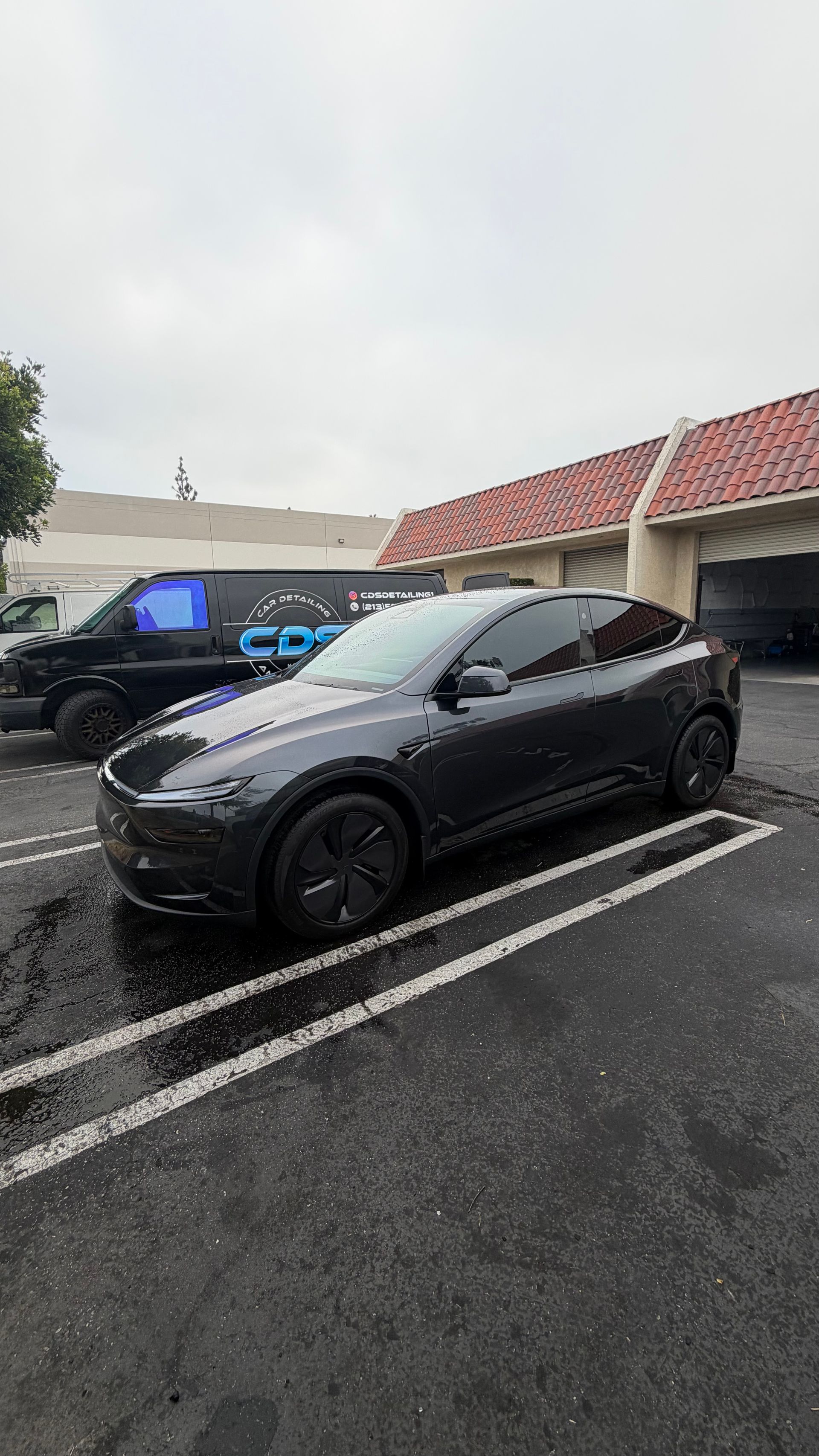 Black Tesla parked in a wet parking lot beside a building with red-tiled roof.