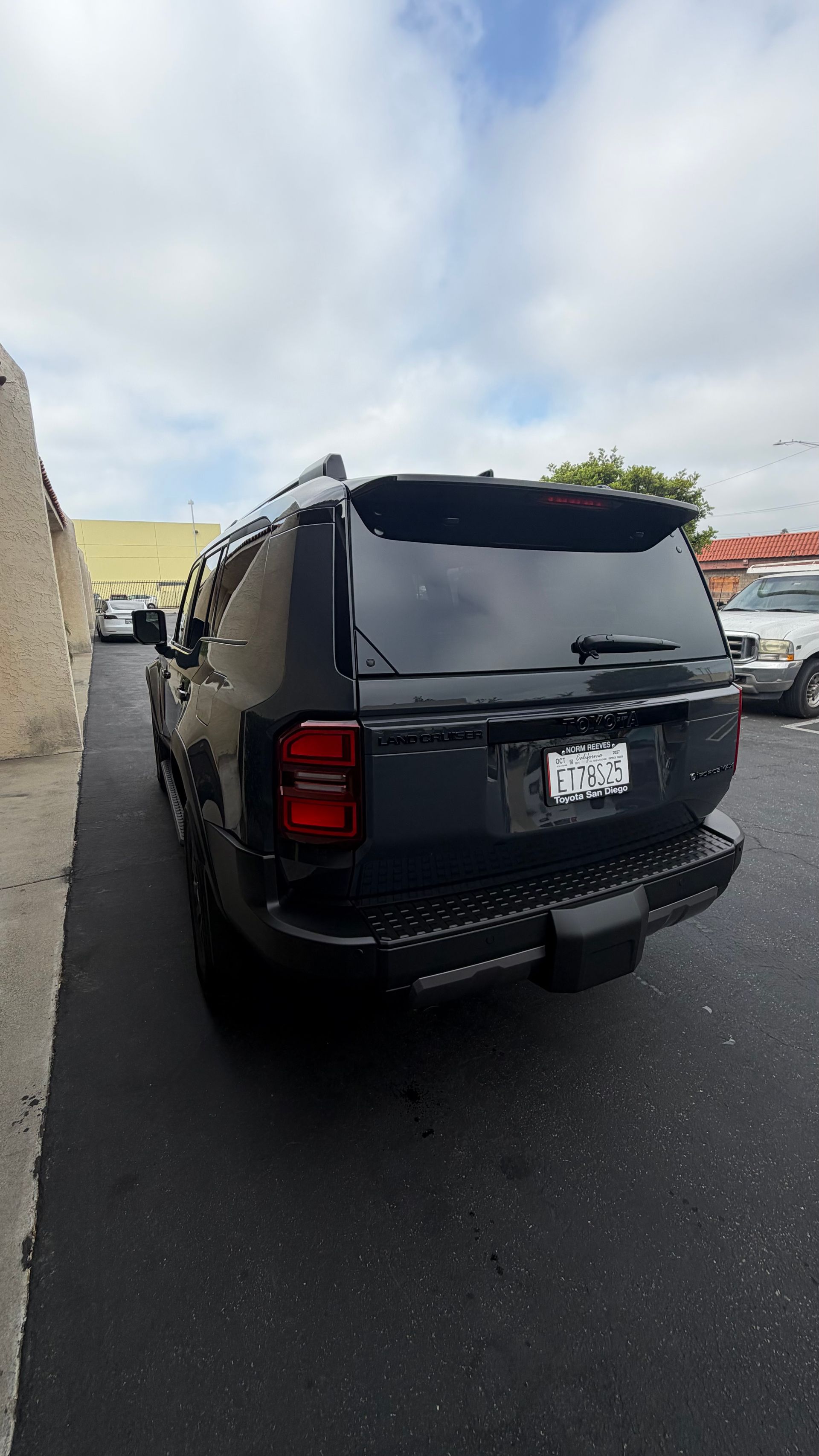 Black SUV parked in a narrow lot beside a wall, viewed from the rear under cloudy skies.