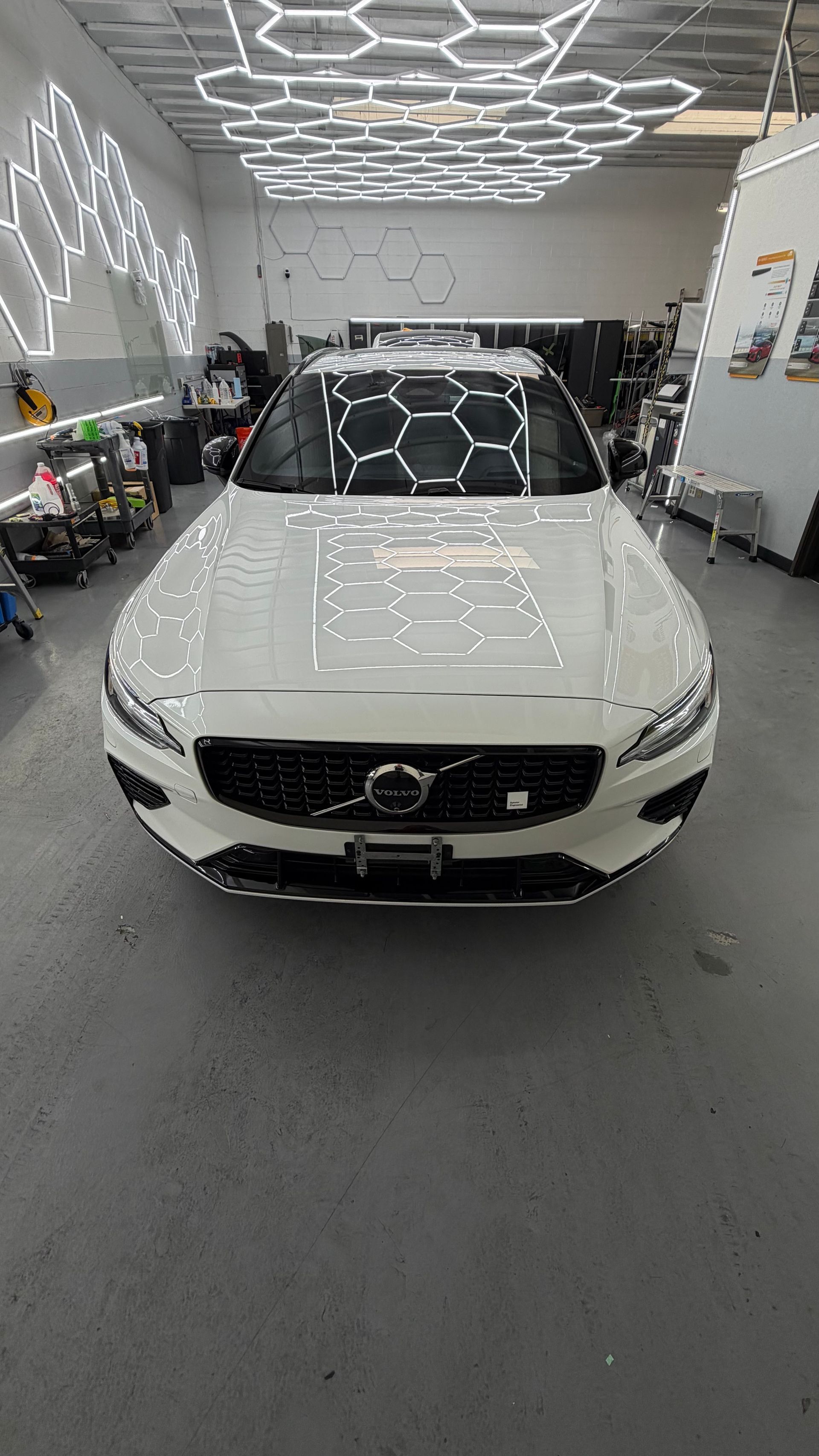 White Volvo SUV in a detailing shop, viewed from the front under bright ceiling lights.