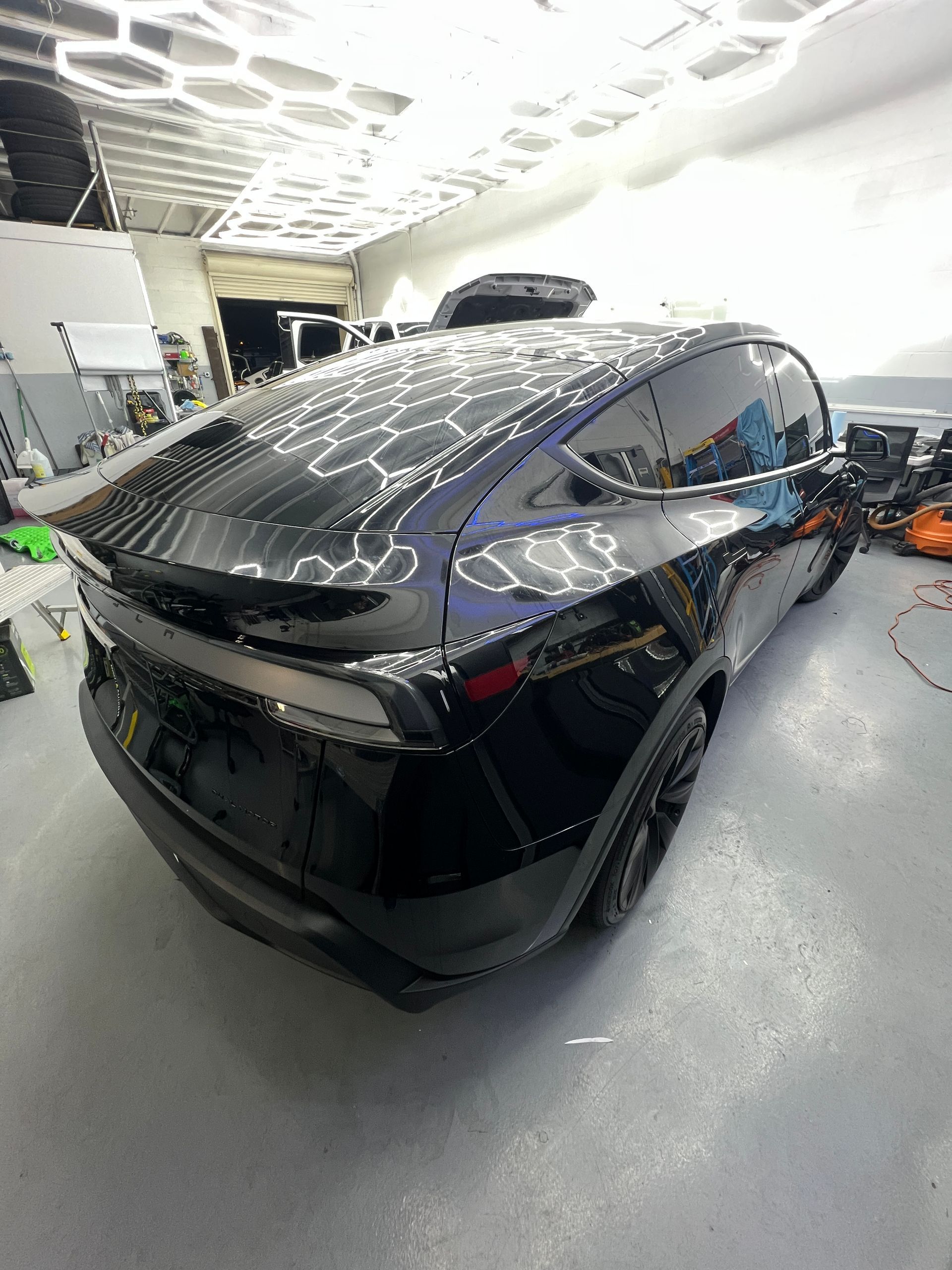Black Tesla SUV in a workshop, rear three-quarter view, with a glossy finish and ceiling light reflections.