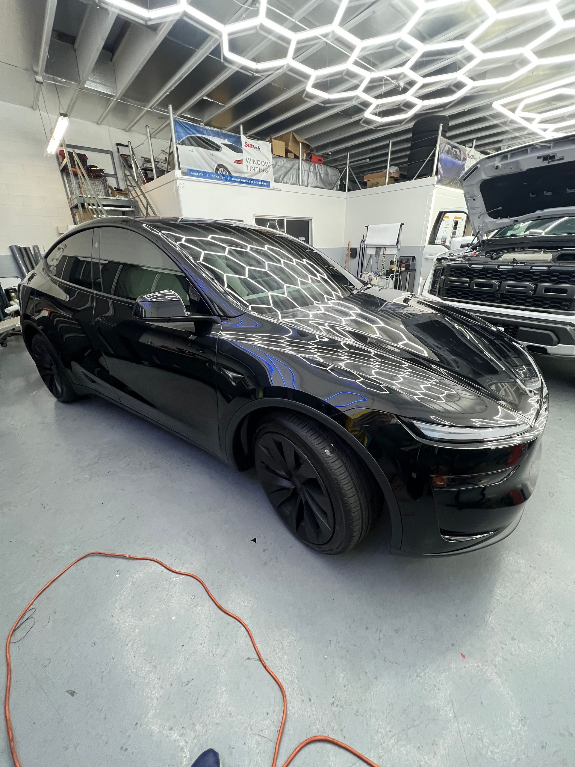 Black Tesla sedan in a bright auto shop with hexagonal ceiling lights reflecting on its body.