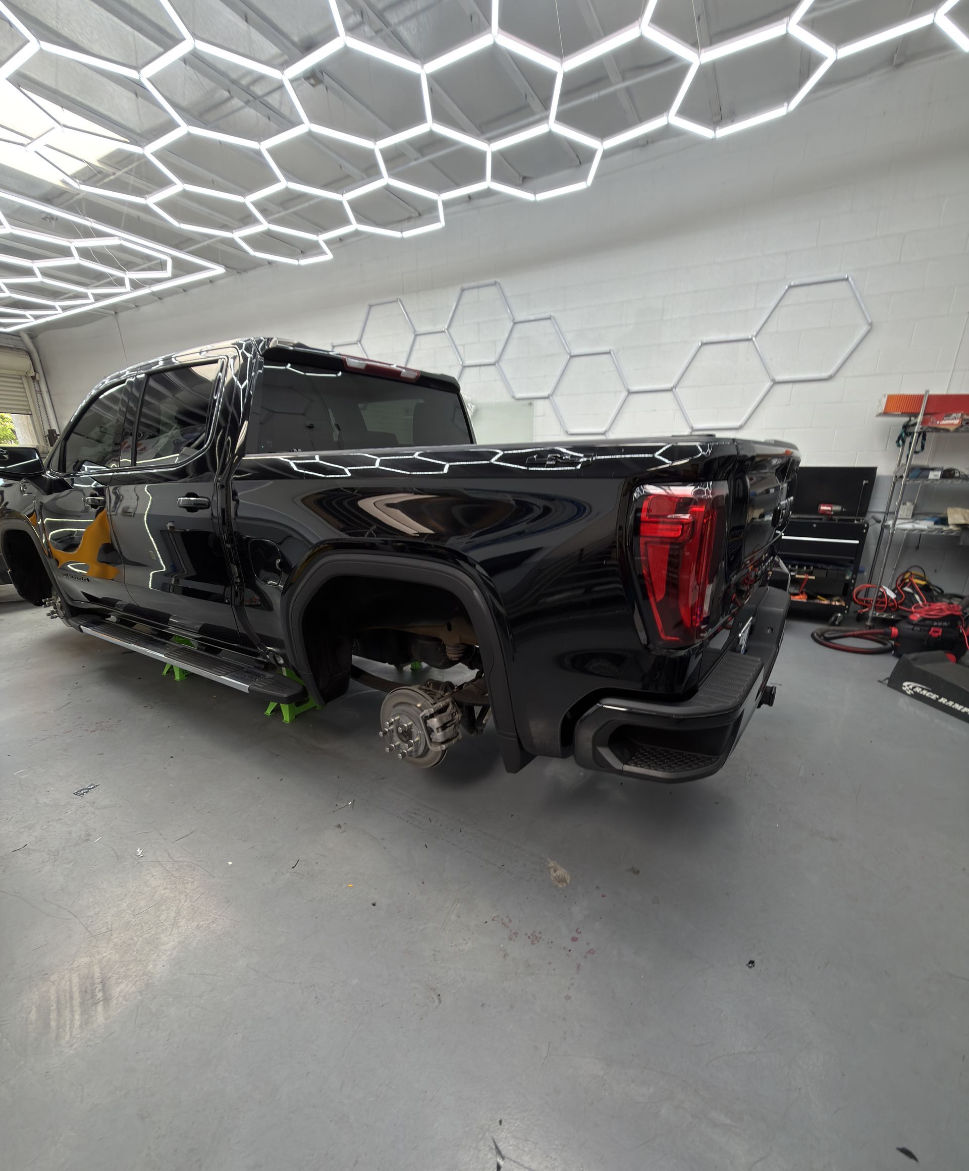 Black pickup truck in a workshop with one rear wheel removed, under hexagon ceiling lights.