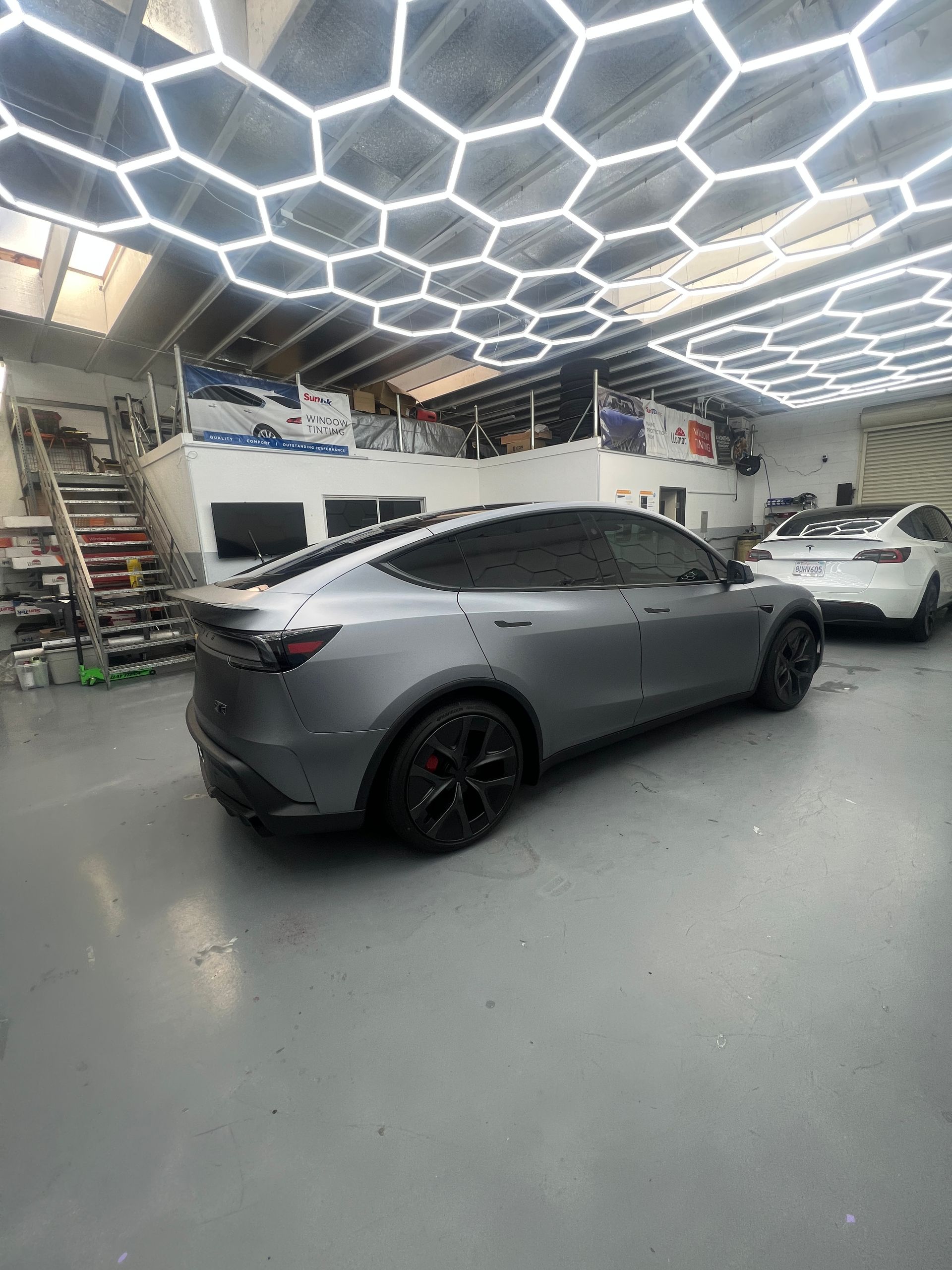 Matte gray sedan in a workshop with hexagon LED ceiling lights and a white car in the background
