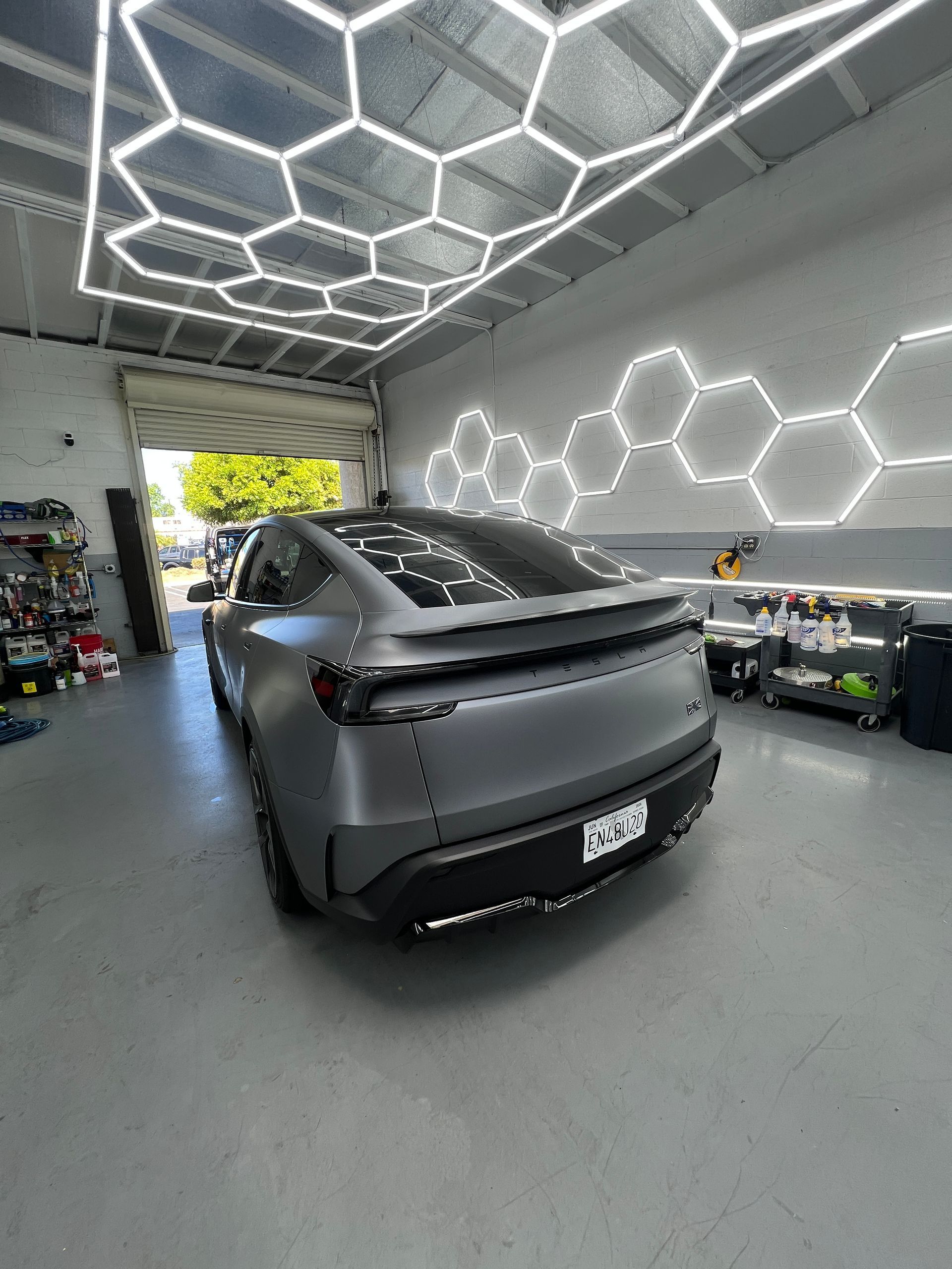 Matte gray sedan in a modern garage with hexagonal LED ceiling and wall lights, rear view.