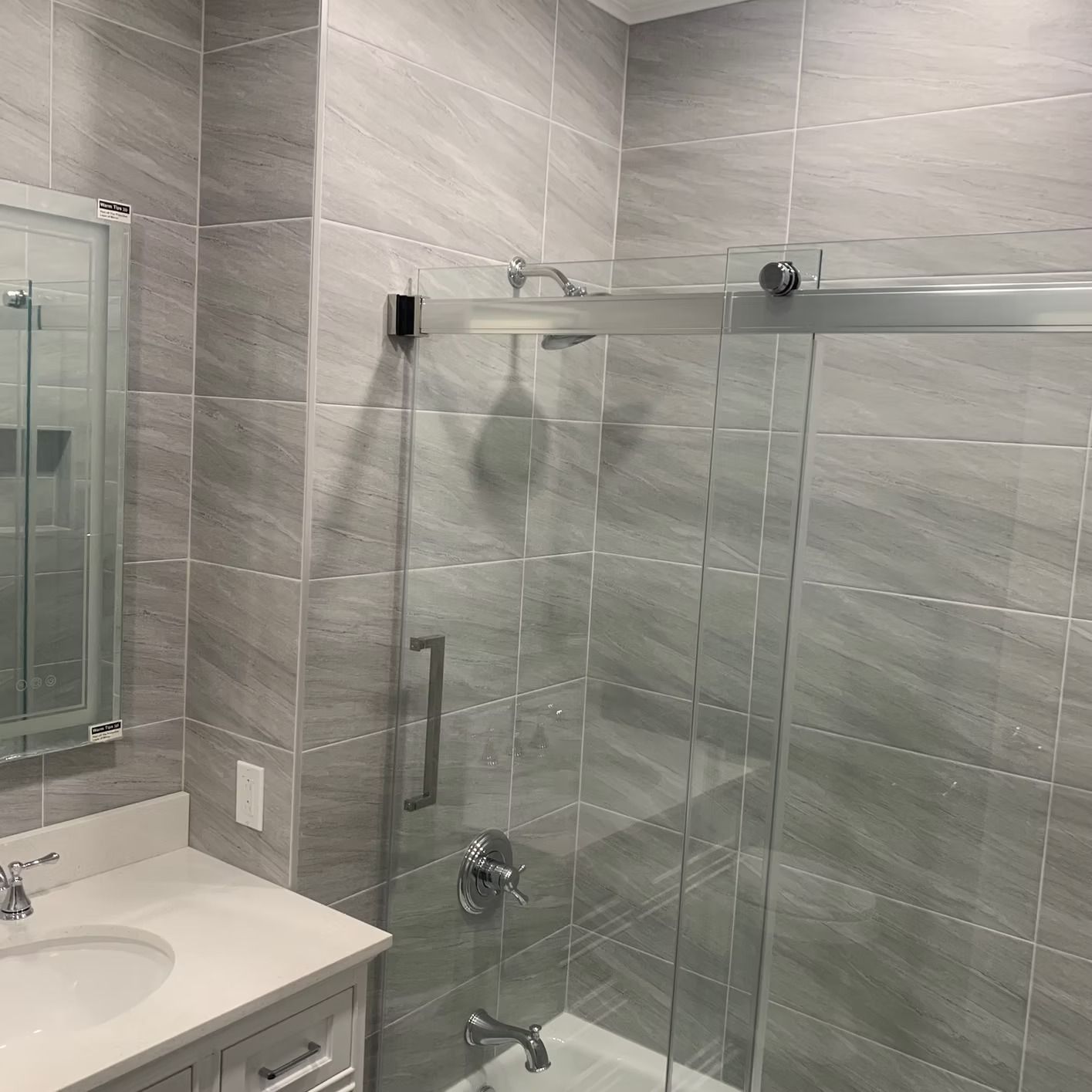 Bathroom with gray tiled walls, glass shower, white countertop, and silver fixtures.