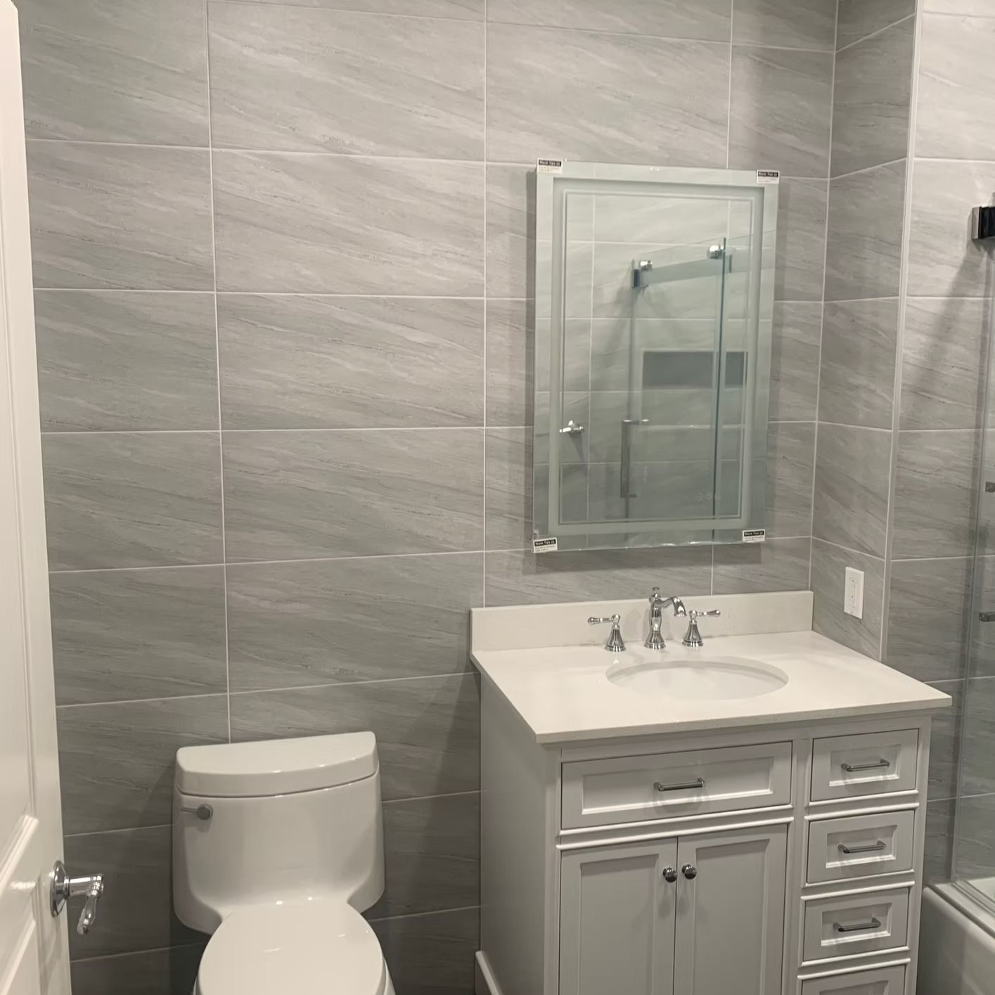 Bathroom with grey tiled walls, white vanity, toilet, and mirror.