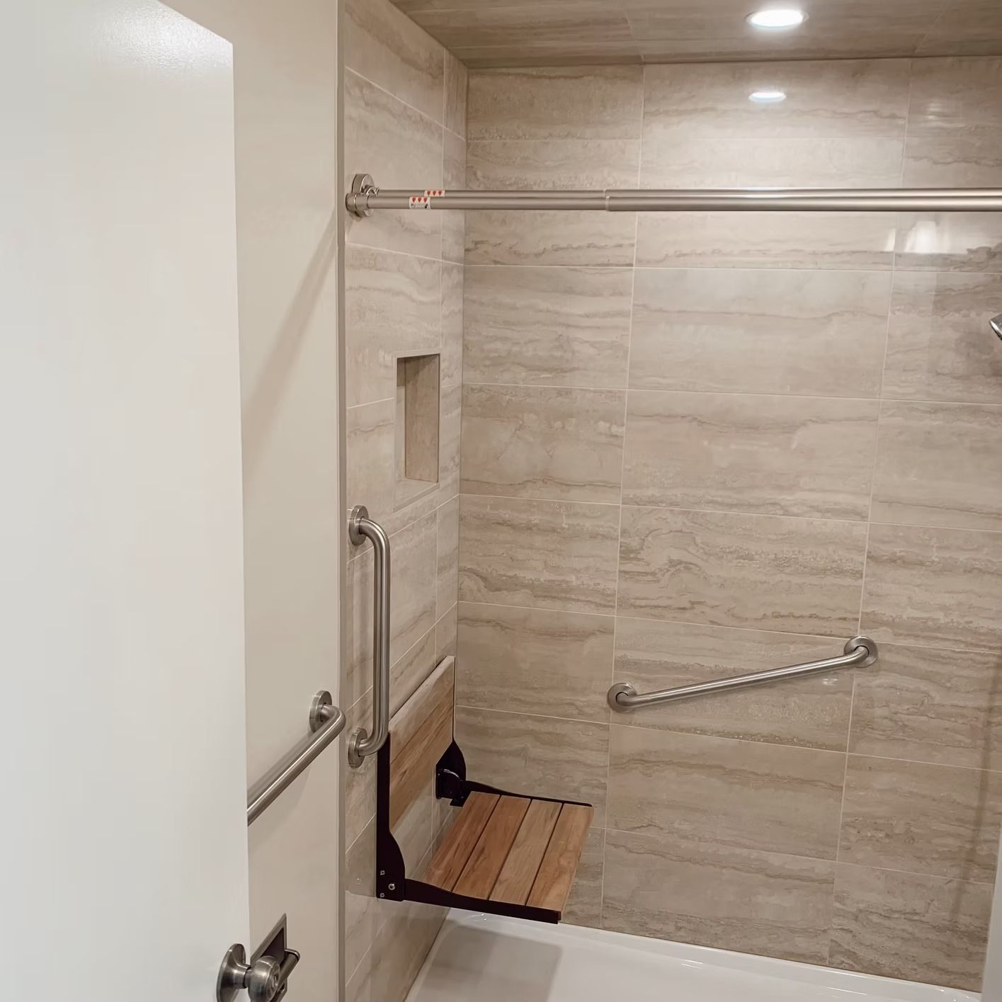 Accessible shower with a folding seat, grab bars, and neutral-colored tile.