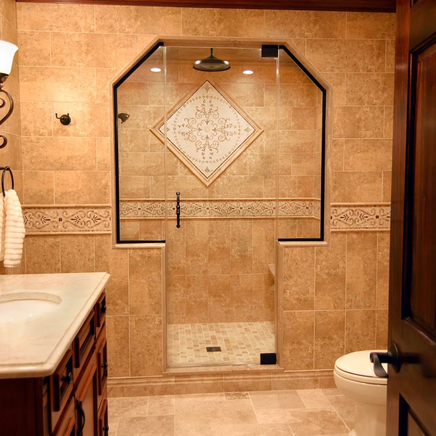 Bathroom with tan tile, glass shower enclosure, ornate tile accent, and wooden vanity.