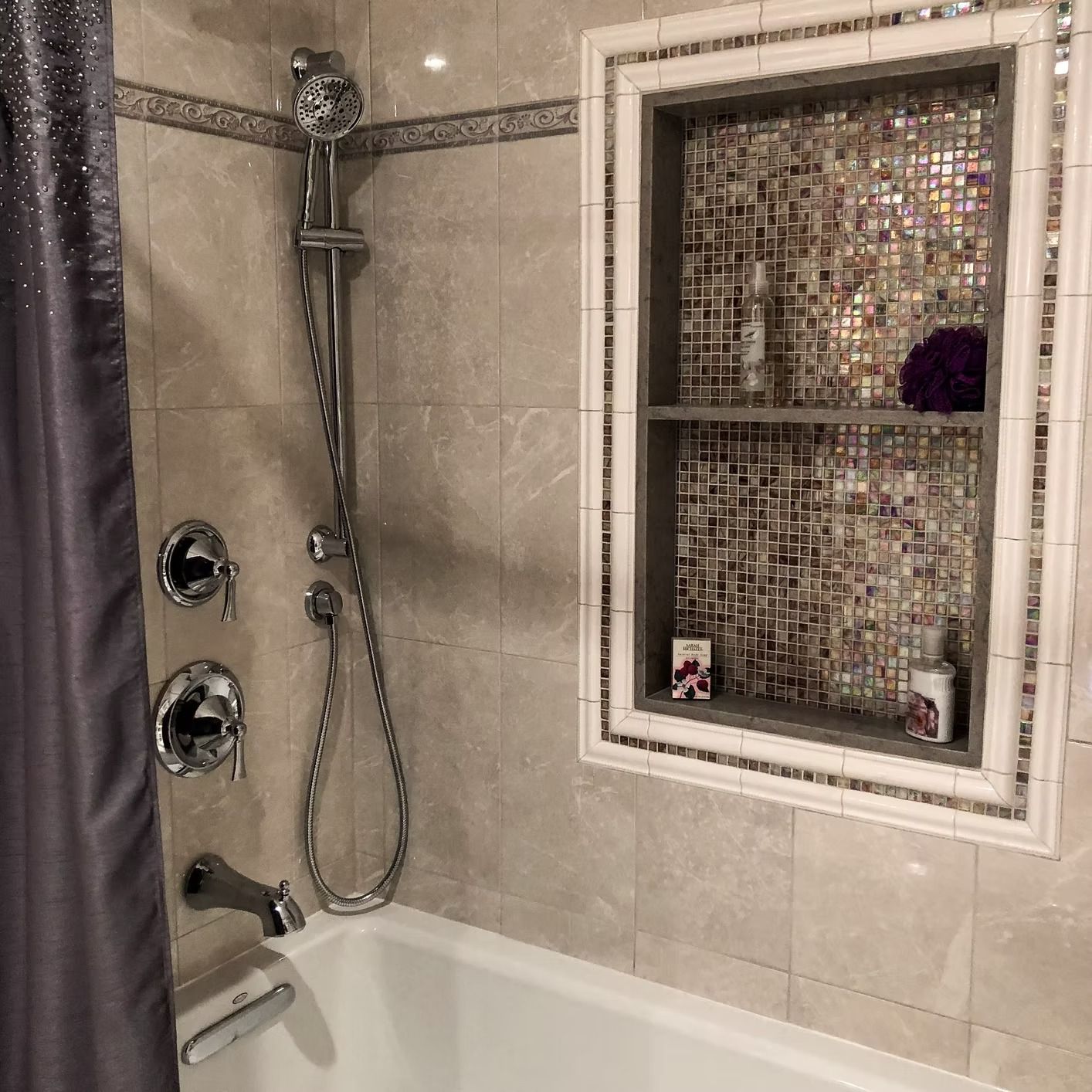 Bathroom with showerhead, fixtures, and built-in shelf. Beige tile walls and white tub.