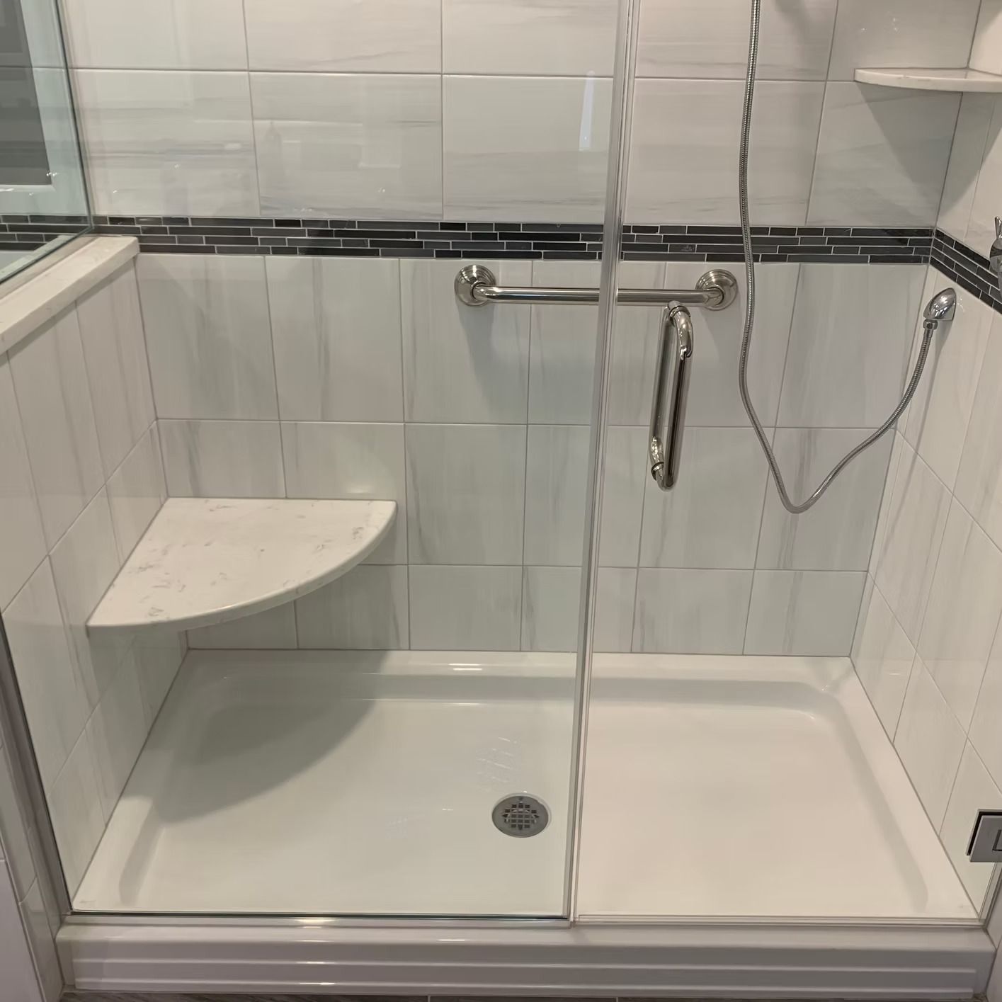 A white tiled shower with a glass door, a seat, and a grab bar.