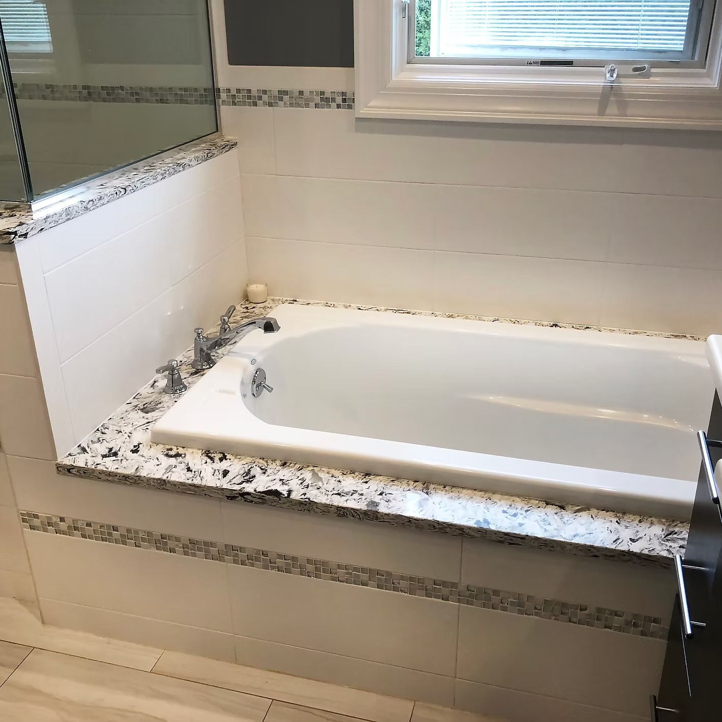 Bathroom with white bathtub, granite countertop, white tiles, and window.