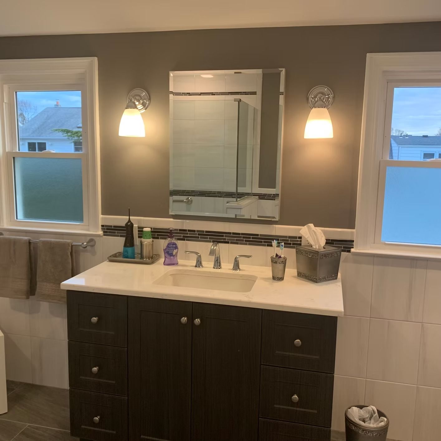 Bathroom with brown vanity, white countertop, mirror, and two sconces. Two windows flank the mirror.