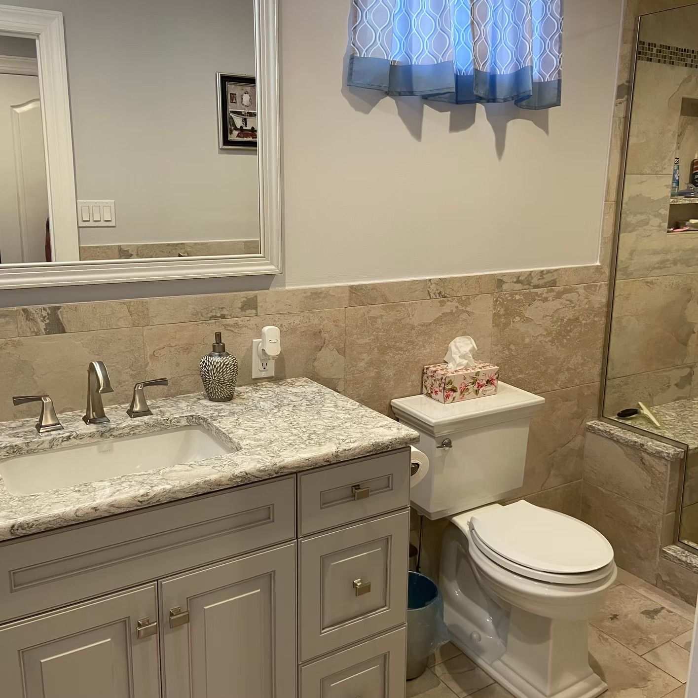 Bathroom with a gray vanity, toilet, and a window with blue curtains. Beige tile on the wall.