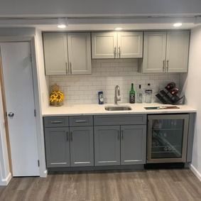 Small gray kitchen with white countertop, cabinets, and a wine fridge. Includes a sink, and liquor bottles.