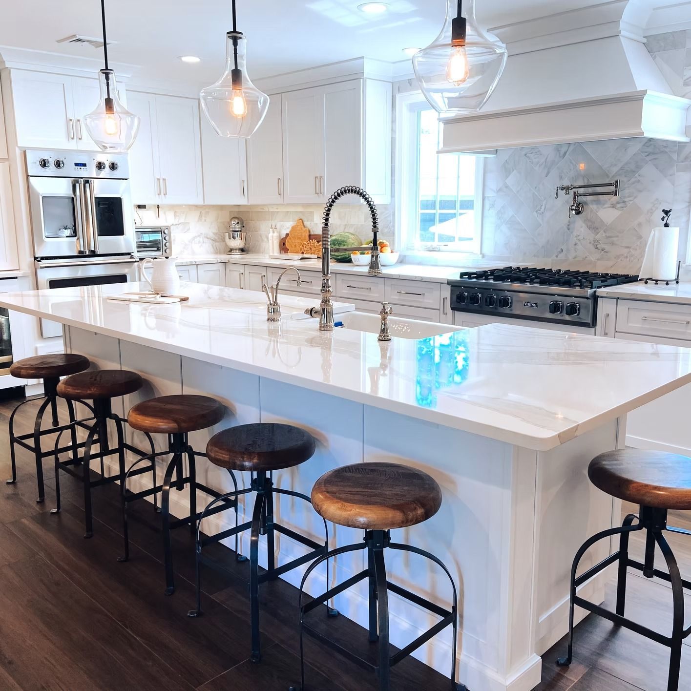 White kitchen with island, six wooden bar stools, and pendant lights.