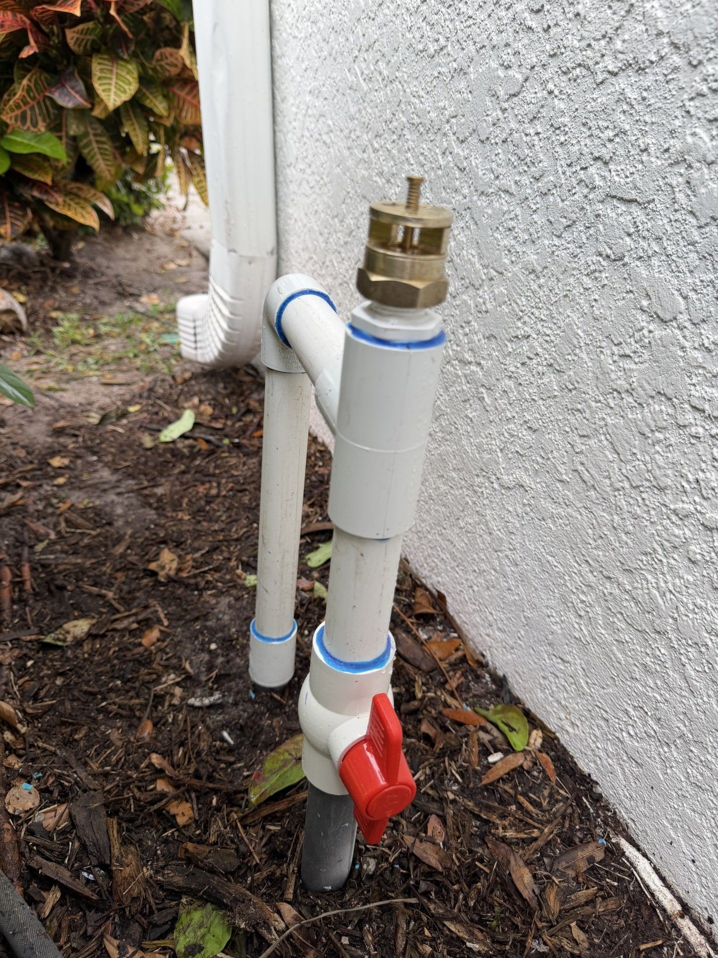 A white PVC pipe assembly with a red shut-off valve and a brass top connector installed against a stucco exterior wall.