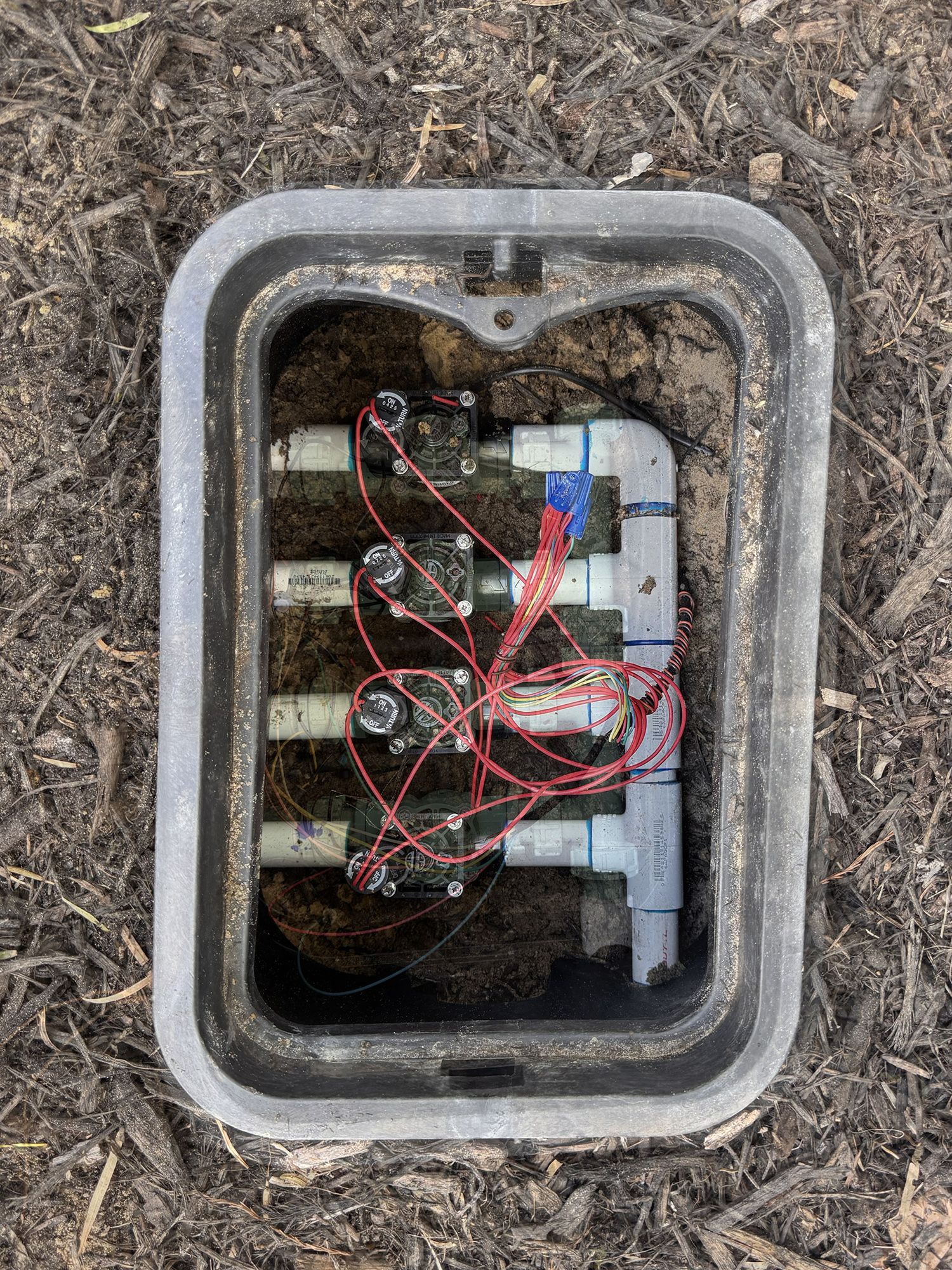 An irrigation valve box containing four green solenoid valves connected to a central pipe, set in mulch.