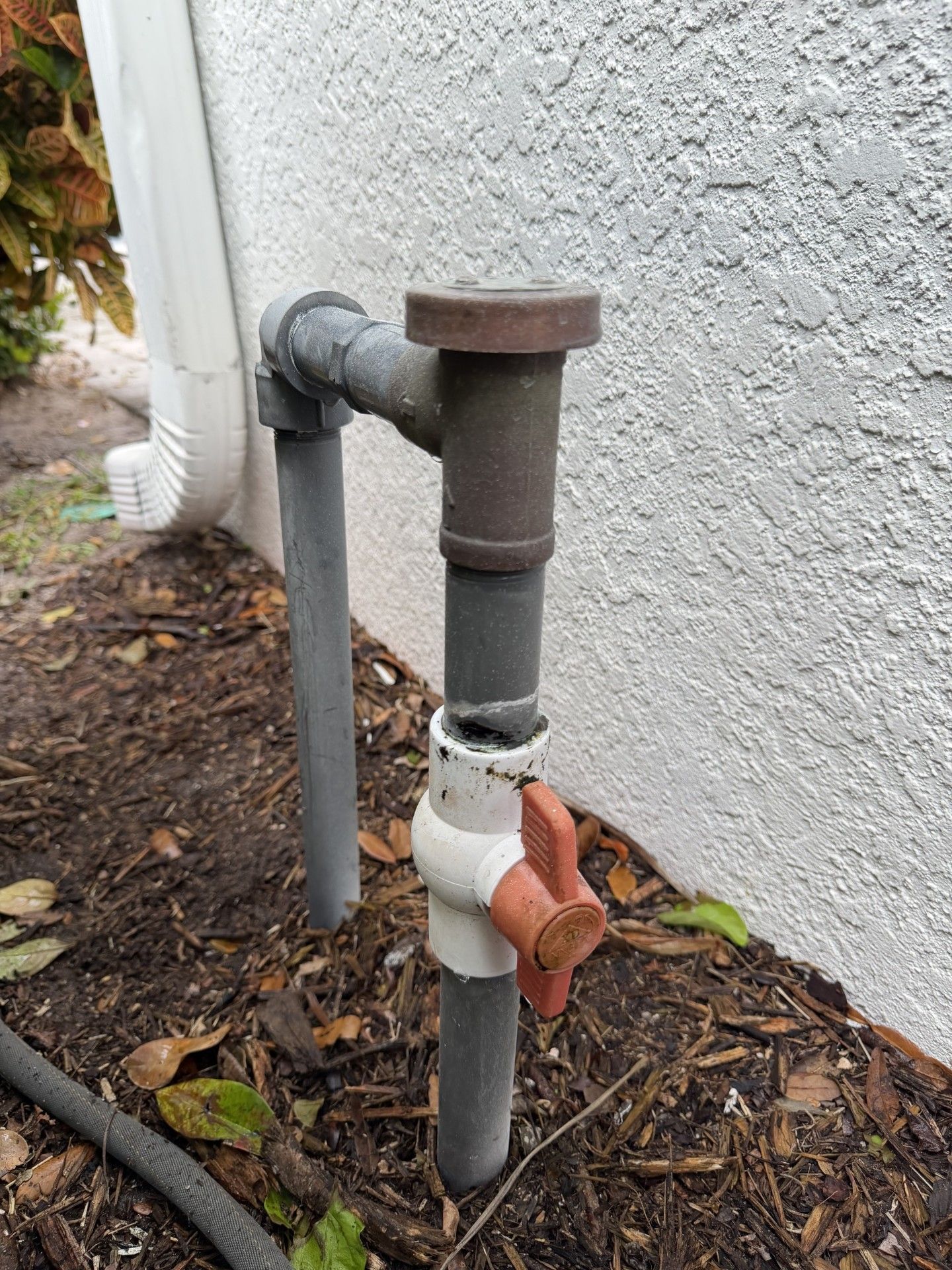 An outdoor water shut-off valve with a red handle connected to metal pipes next to a house wall.