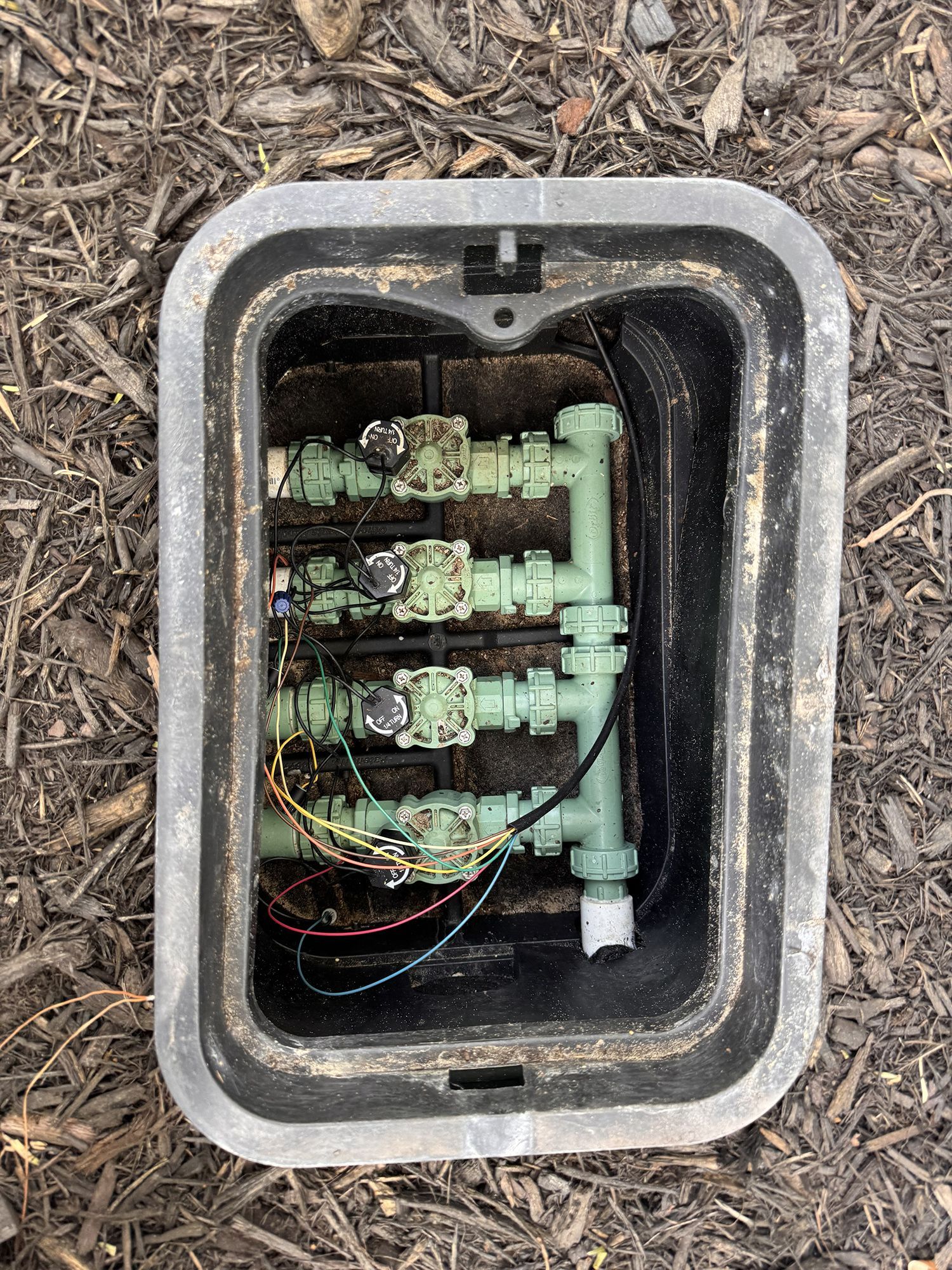 An irrigation valve box in the ground containing four valves connected by white PVC pipes and red electrical wiring.
