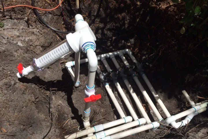 A PVC irrigation system manifold with a clear cylindrical filter and red ball valves, partially buried in dirt.