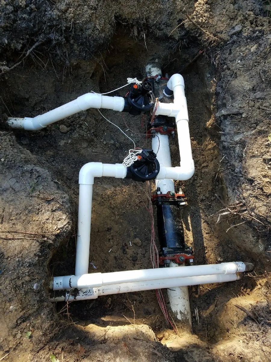 An underground irrigation manifold with white PVC pipes, black valves, and control wires, installed in a dirt trench.