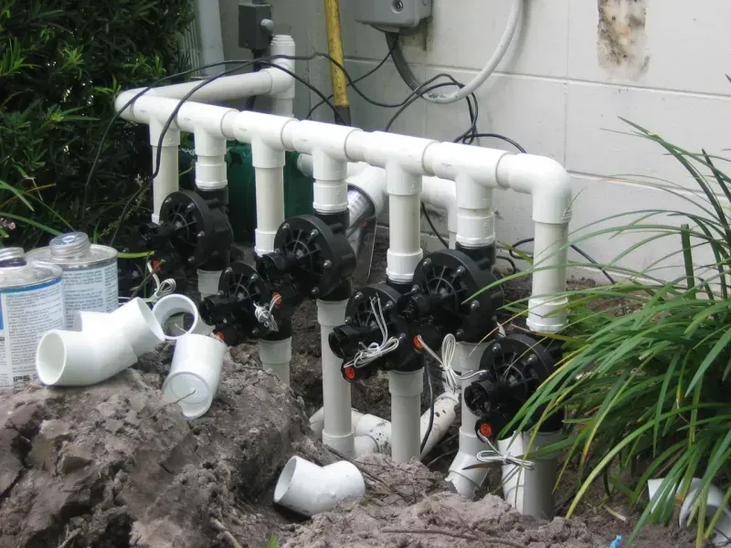 A row of four irrigation valves mounted on white PVC pipes next to an exterior wall with loose fittings on the ground.