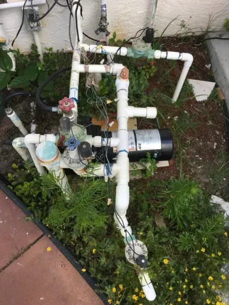 Outdoor white PVC plumbing and irrigation pump system with wiring, partially obscured by weeds and vegetation.