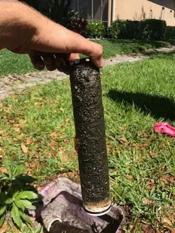 A person holds a heavily soiled, dark brown cylindrical water filter outdoors over a utility box.