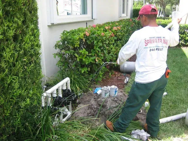 A worker in a white long-sleeved shirt repairs a PVC sprinkler manifold system outside a home.