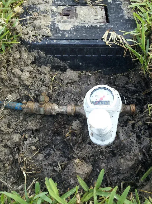 A residential water meter and shut-off valve installed in the dirt ground next to a black utility box lid.