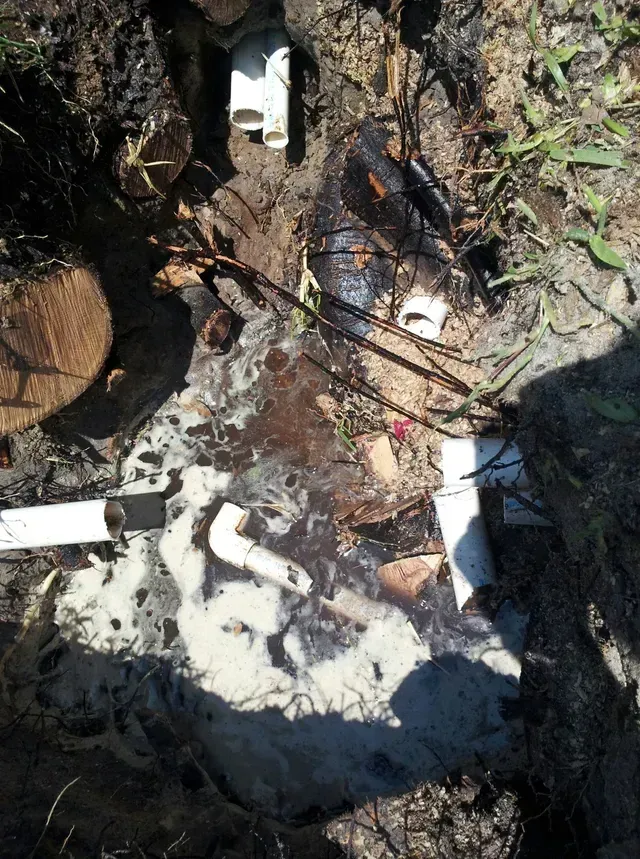 A ground-level view showing broken PVC pipes leaking a foamy white substance into a muddy dirt hole.
