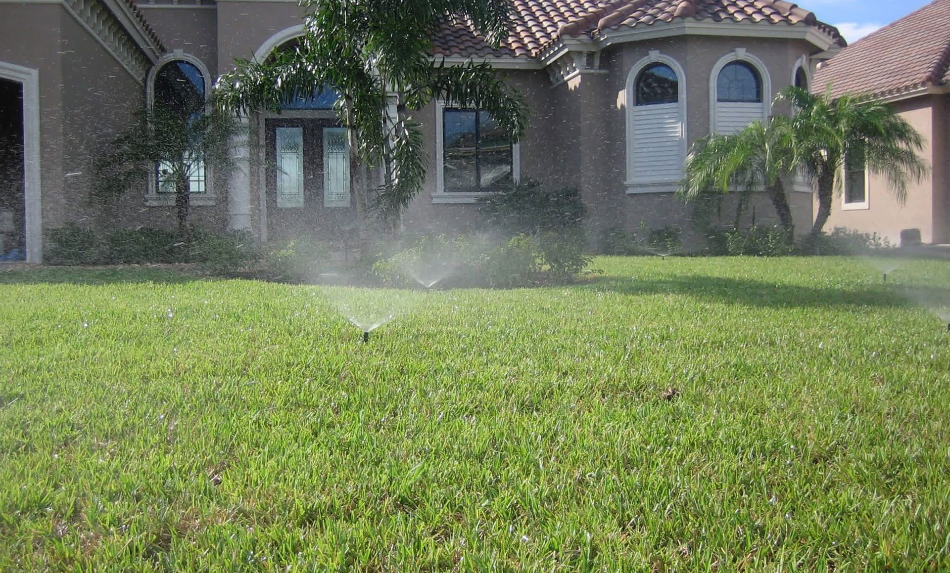 A residential lawn with green grass being watered by an irrigation sprinkler in front of a house.