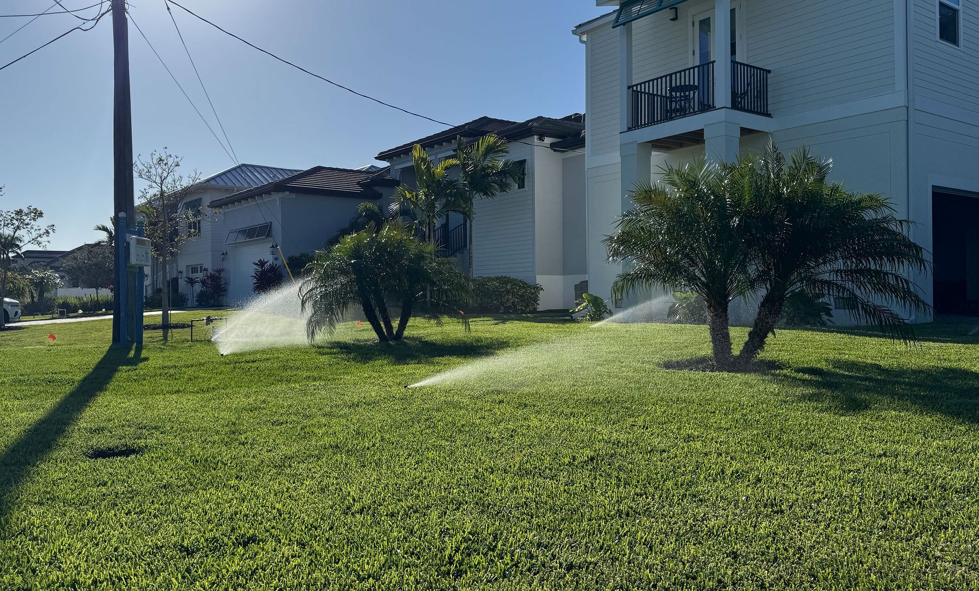 Residential lawn sprinklers spray water over green grass in front of houses under a clear blue sky.